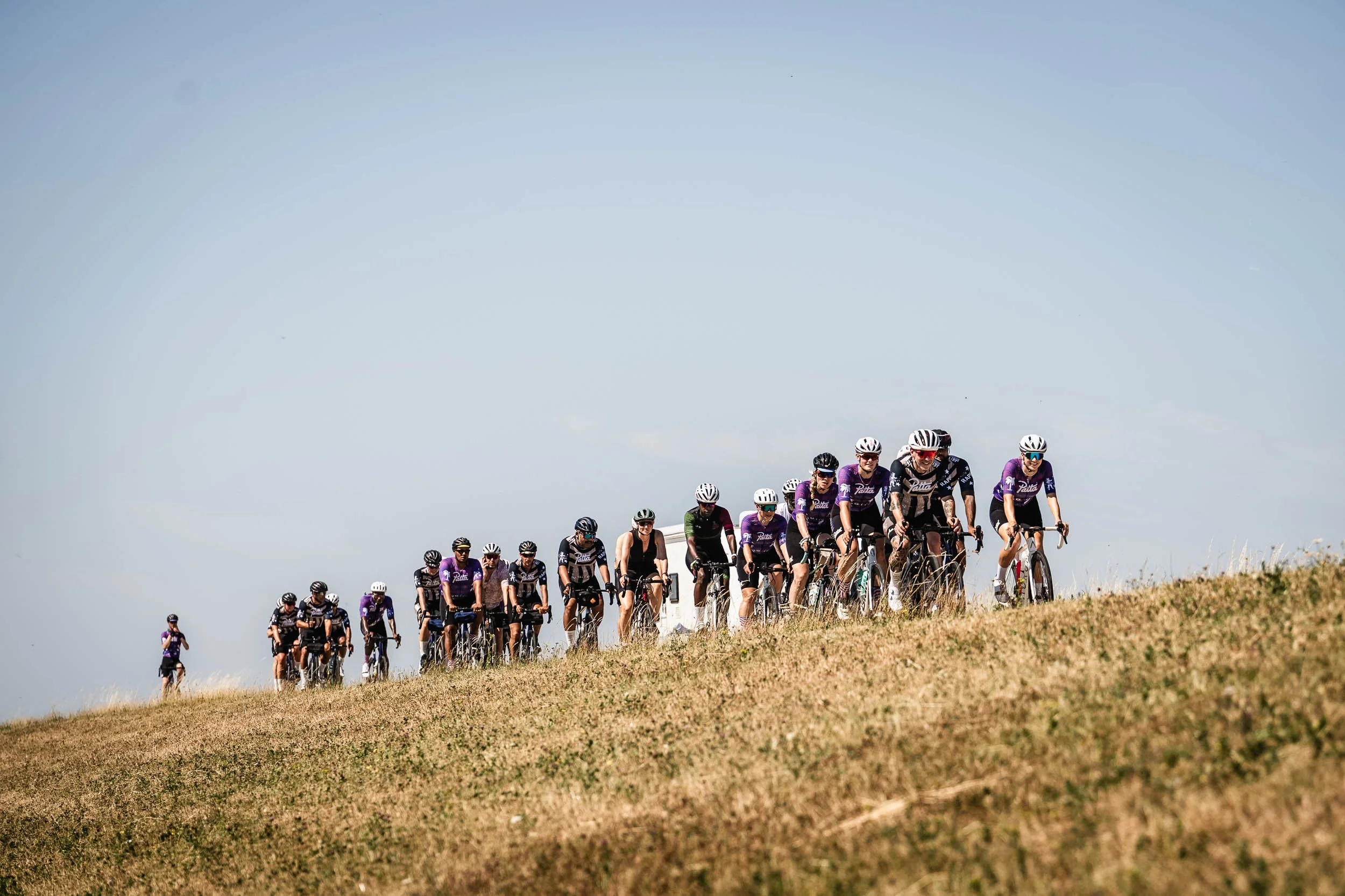 A group of cyclists riding on a grassy hill during daytime, wearing helmets and cycling gear. (Patta.nl Rapha)