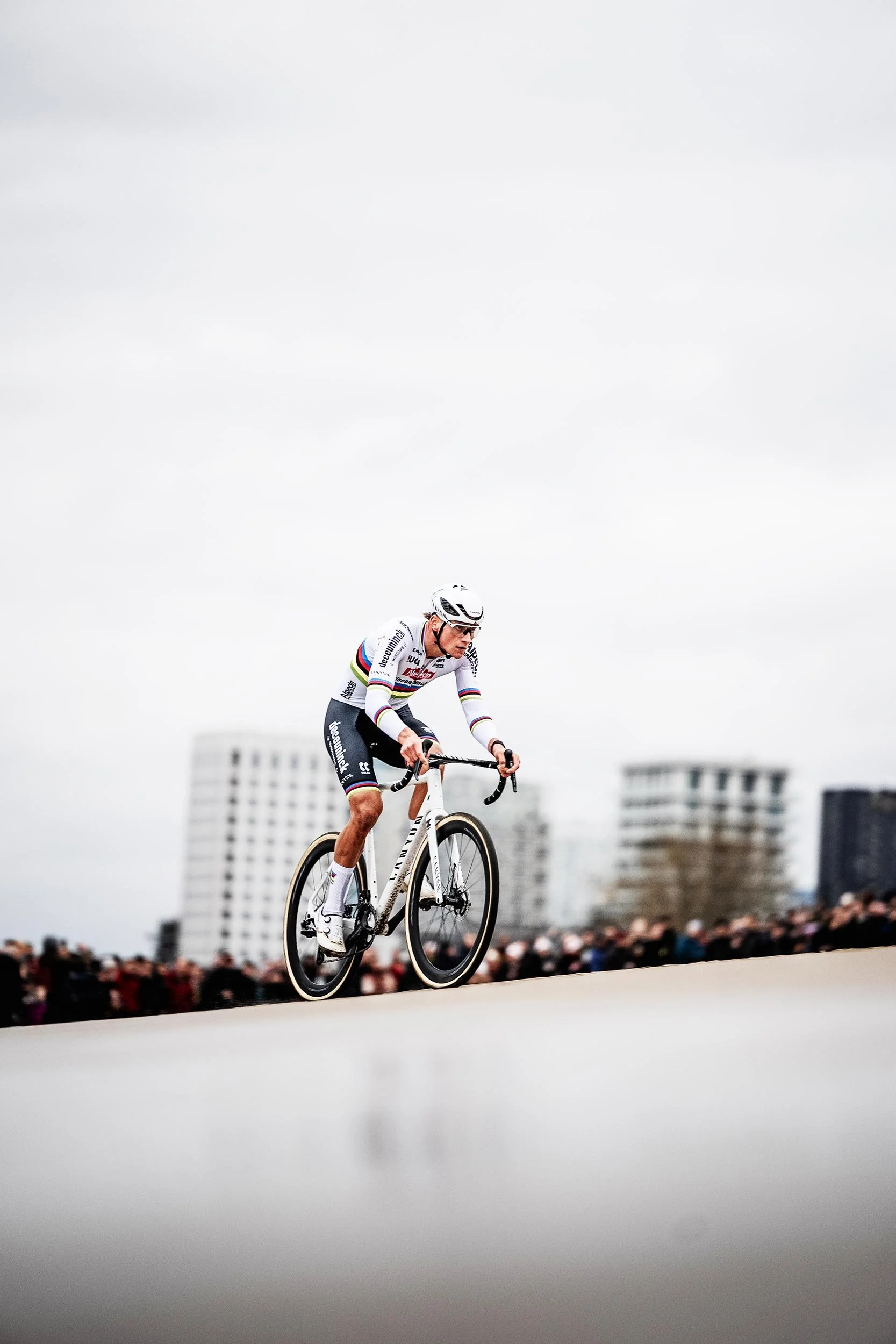 A cyclist in racing gear, wearing a rainbow-striped jersey and helmet, riding a white racing bicycle on a road with an audience and tall buildings in the background.