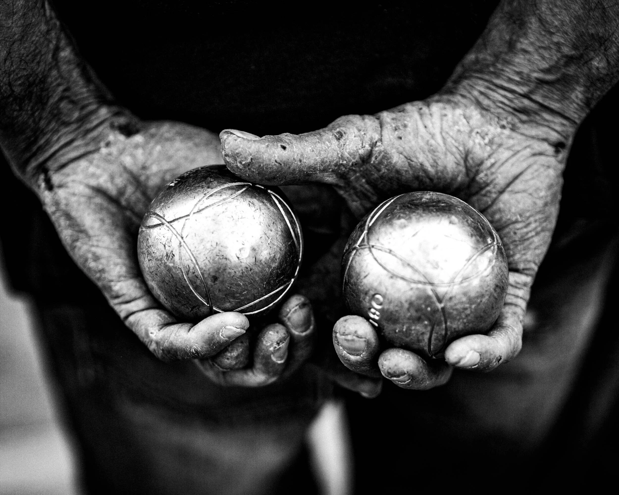 Close-up of elderly person's hands holding two worn metallic petanque balls, with visible veins and aged skin.