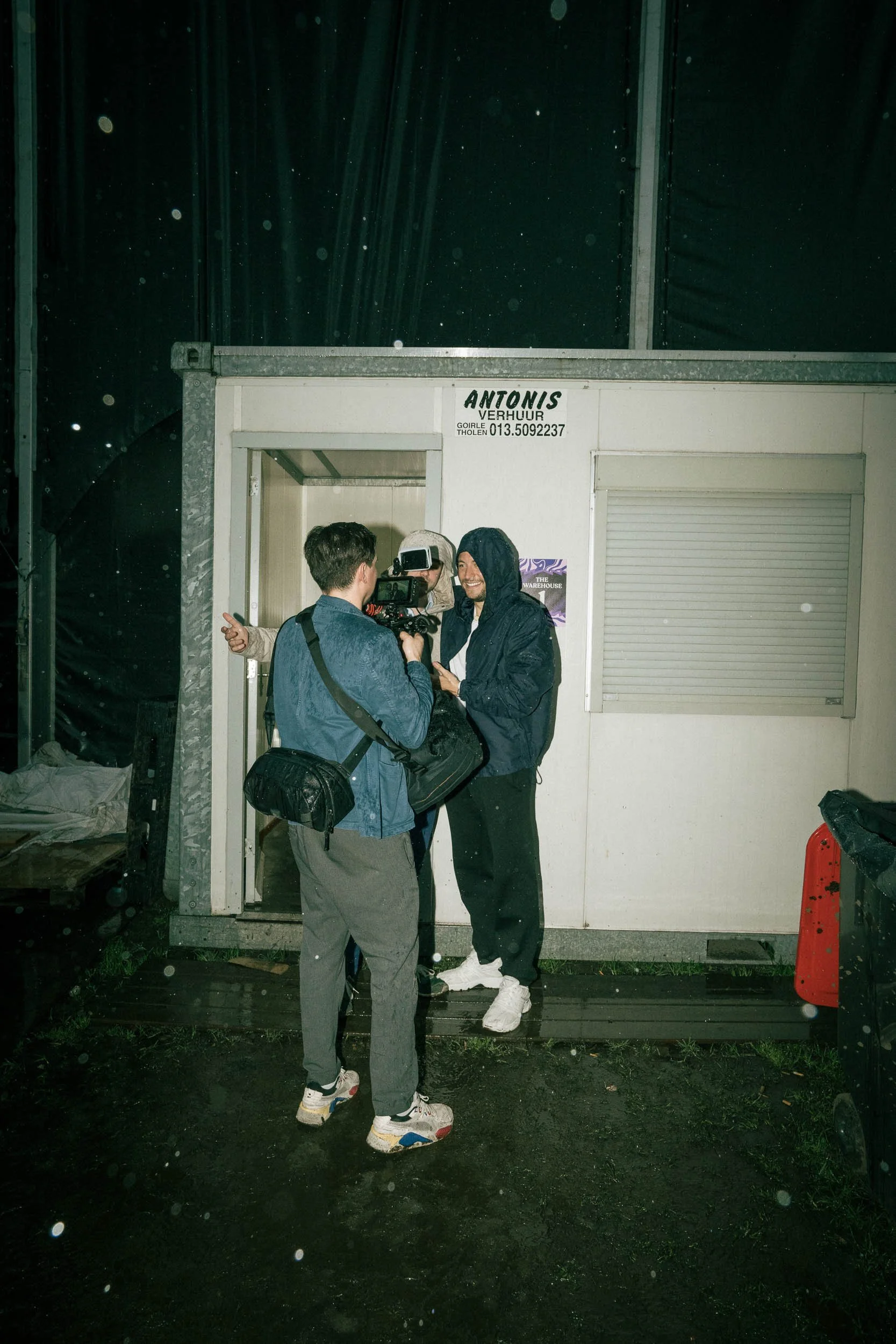 Two men are filming a person in casual clothing outside a small building with a sign that reads 'ANTONIS VERHUIR'. It is raining at night.  (Viktor Verhulst)