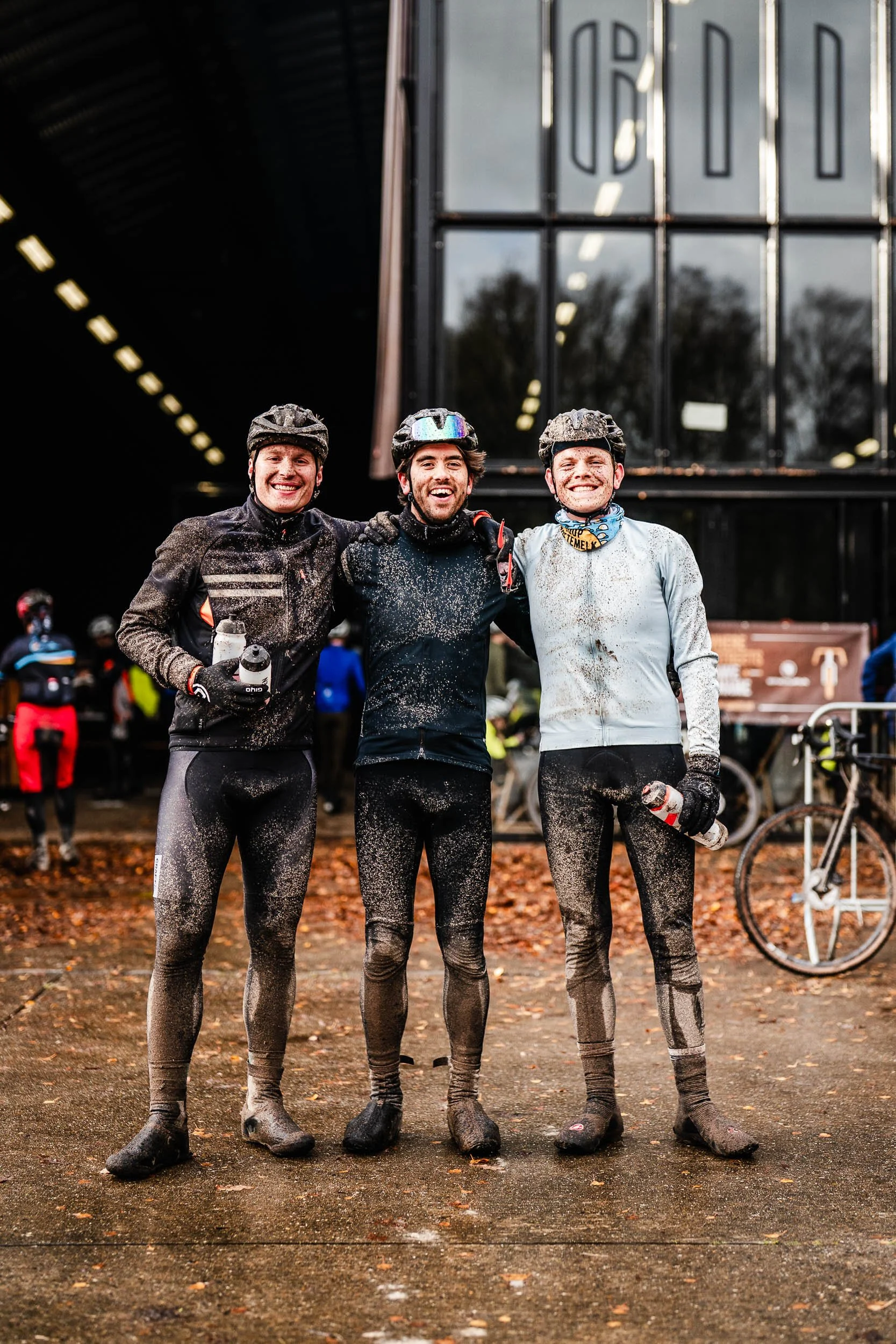 Three male cyclists smiling and posing together after a muddy race, holding water bottles, with a large building in the background.