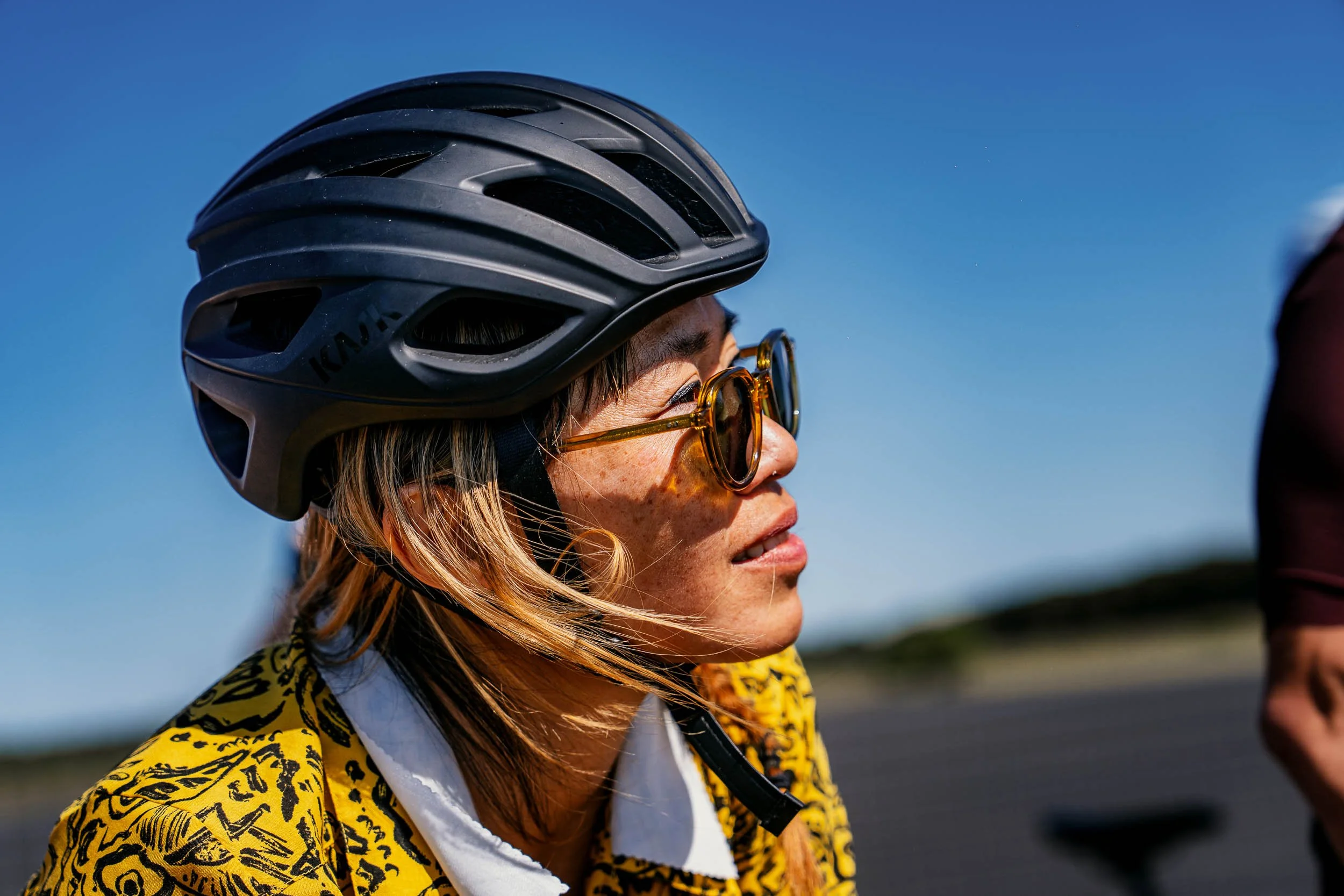 Close-up of a woman wearing a black bike helmet, yellow patterned jacket, and sunglasses, with her face turned slightly to the side against a clear blue sky.