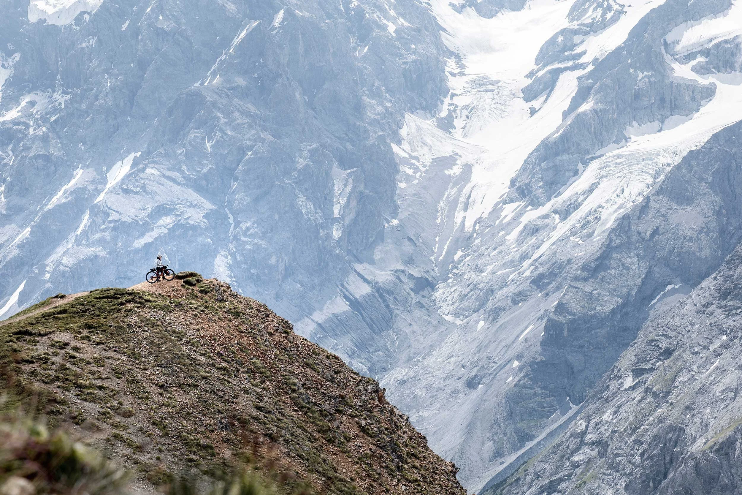 A person with a bicycle standing on a grassy hilltop with snow-capped mountains in the background.