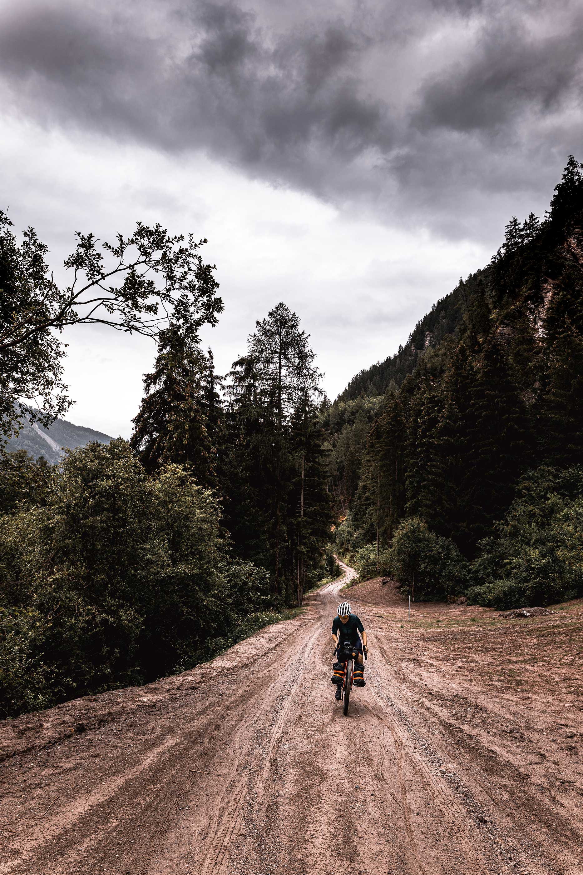 A person riding a bicycle on a dirt trail through a forested mountain landscape under a cloudy sky.