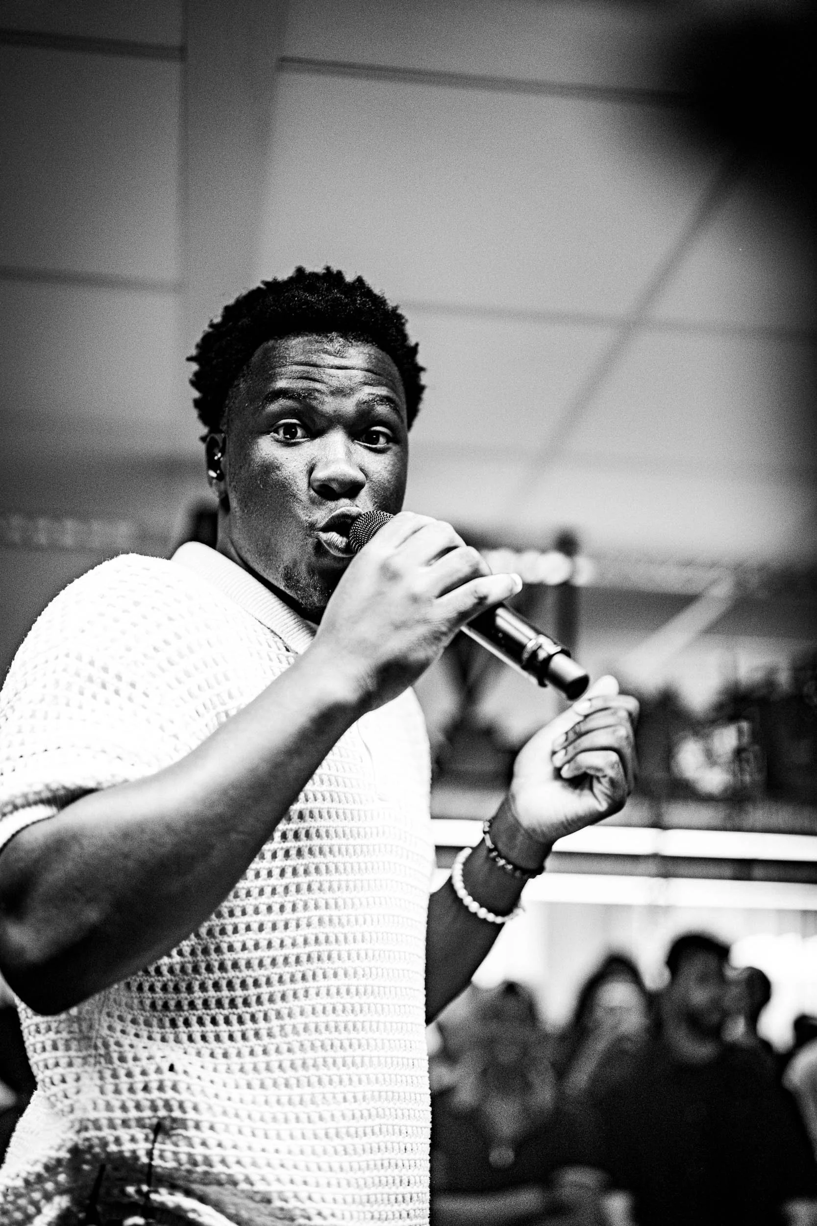 A person with short curly hair holding a microphone, looking directly at the camera, with a surprised or expressive facial expression, in a room with a ceiling grid and blurred background of people.  (Viktor Verhulst)