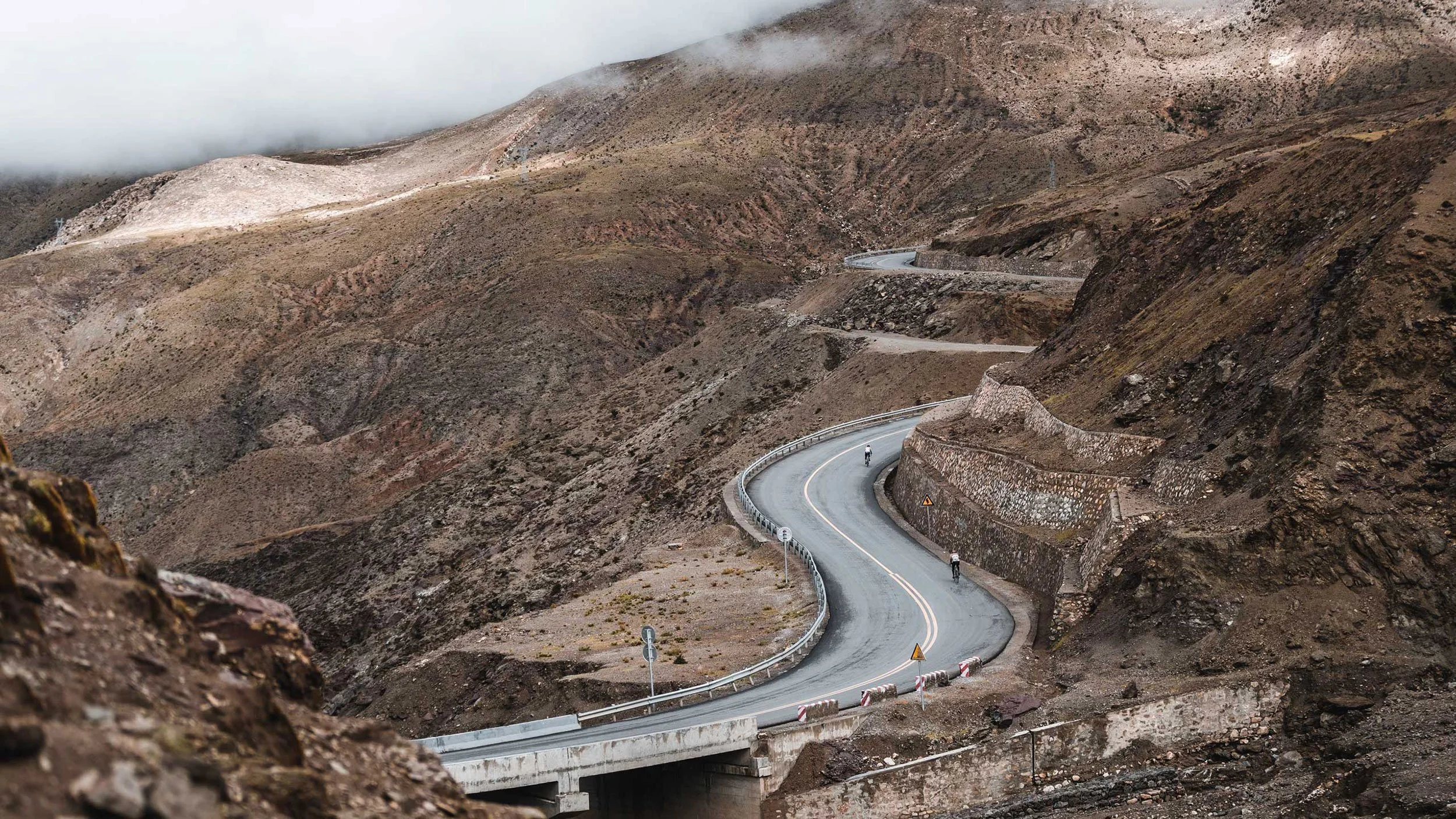 A winding mountain road with barriers on the sides, surrounded by barren rocky terrain and fog-covered peaks in the distance. Two cyclists are riding along the road.