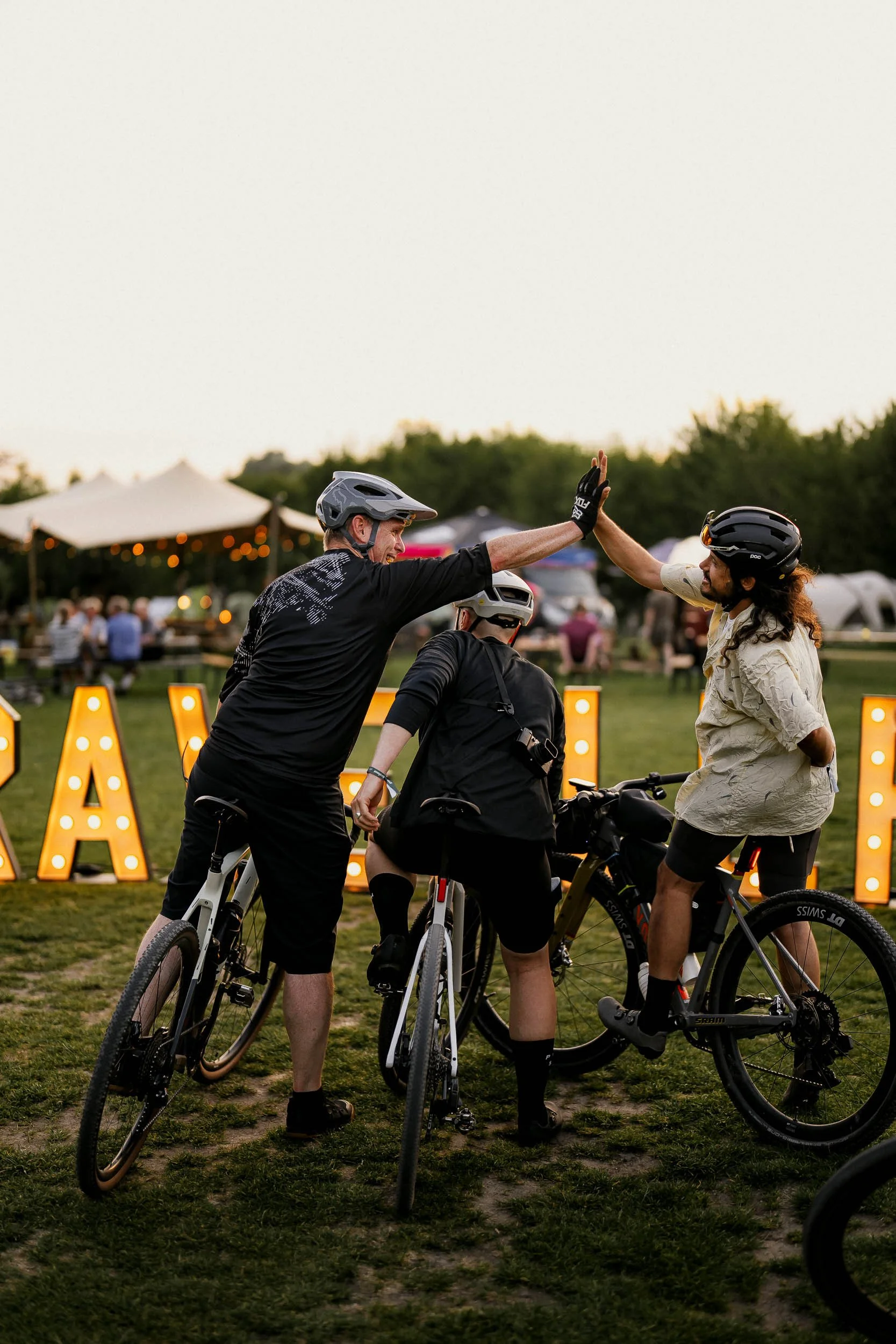 Three people on bicycles giving each other high fives at an outdoor event during the evening, with tents and illuminated signs in the background.