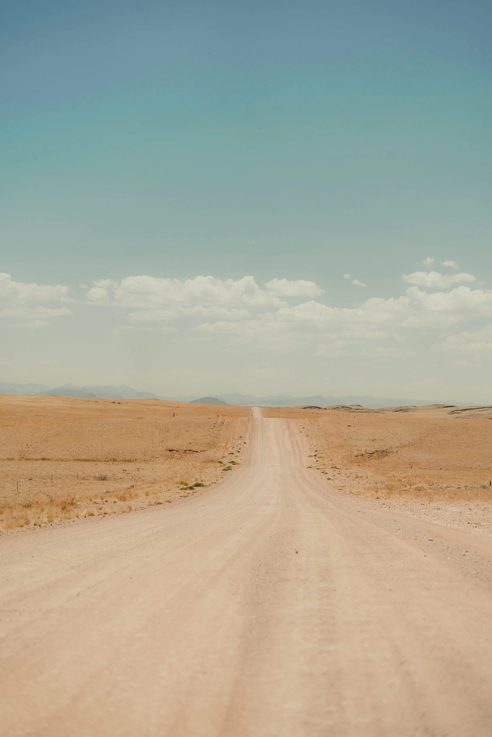 A straight dirt road extends through a vast, open desert landscape with sandy terrain and minimal vegetation, under a blue sky with scattered clouds.