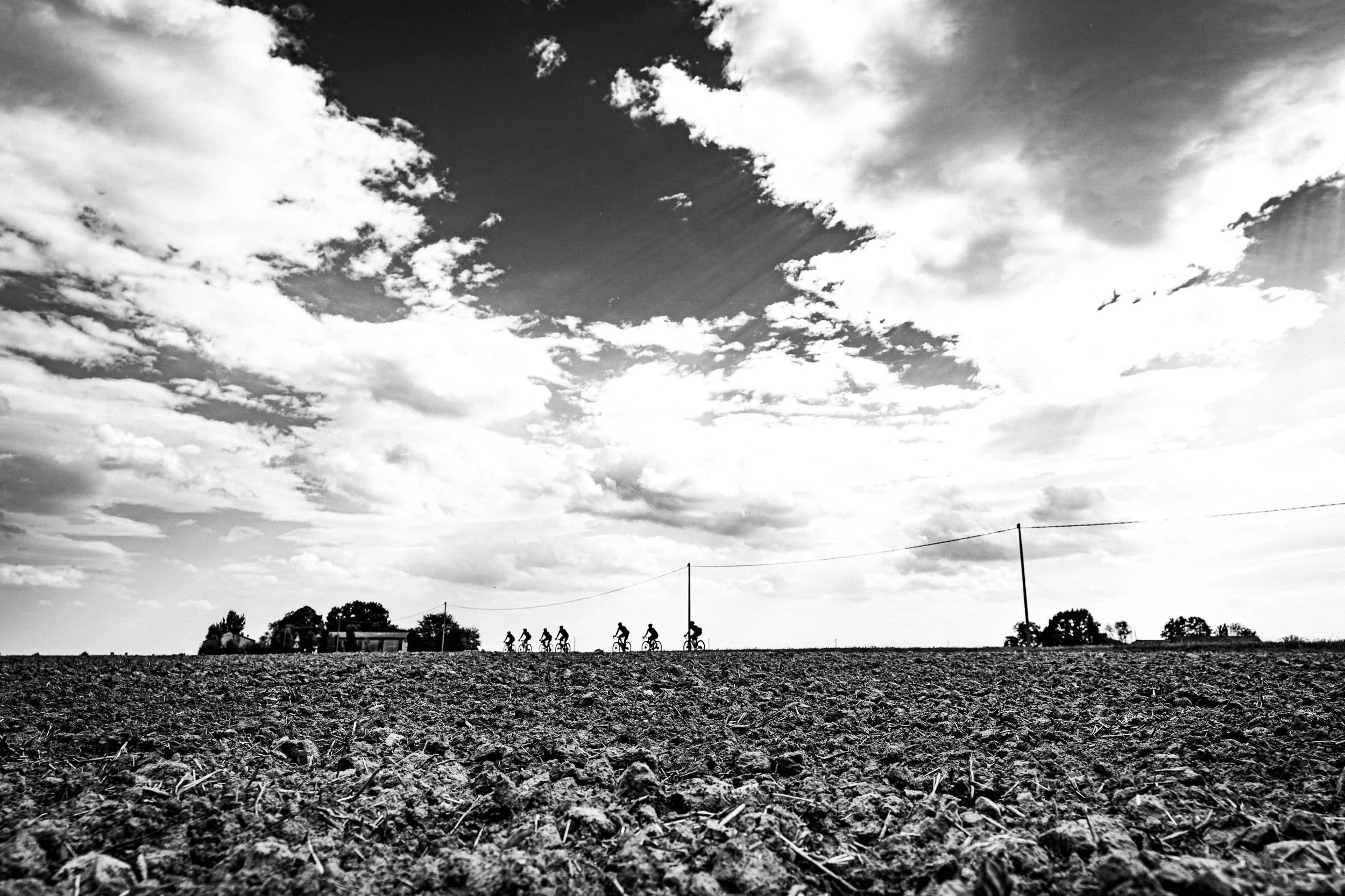 A group of six cyclists riding on a dirt road under a cloudy sky, with a few poles and trees in the distance.