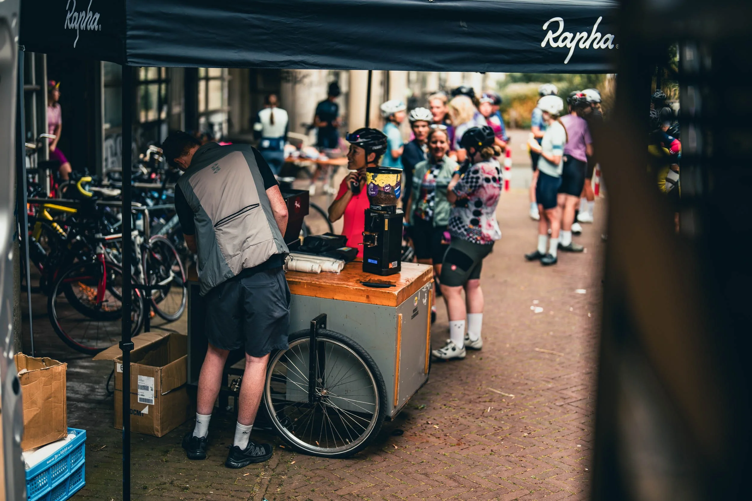 A crowd of cyclists gathered at a registration or check-in area for a bike event. Several people are wearing cycling gear and helmets, some are talking, and bikes are visible in the background. A man is at a table under a blue canopy.