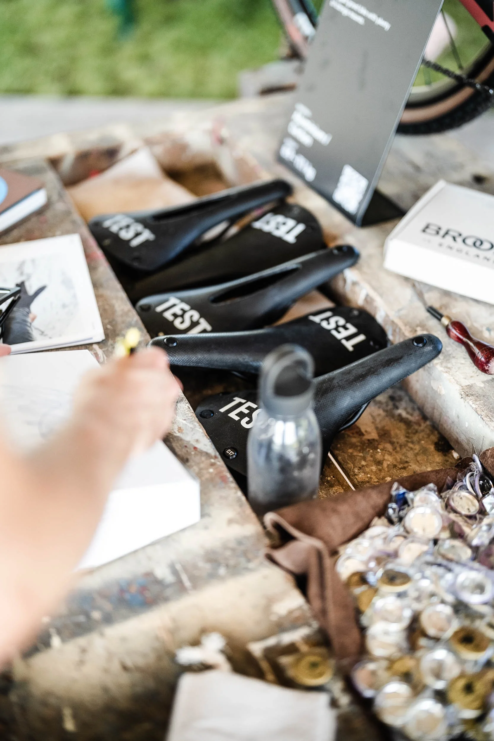 A row of black bicycle seats labeled 'TEST' on a rustic counter, with a hand holding a small tool, a water bottle, and various small items like coins and papers around them.