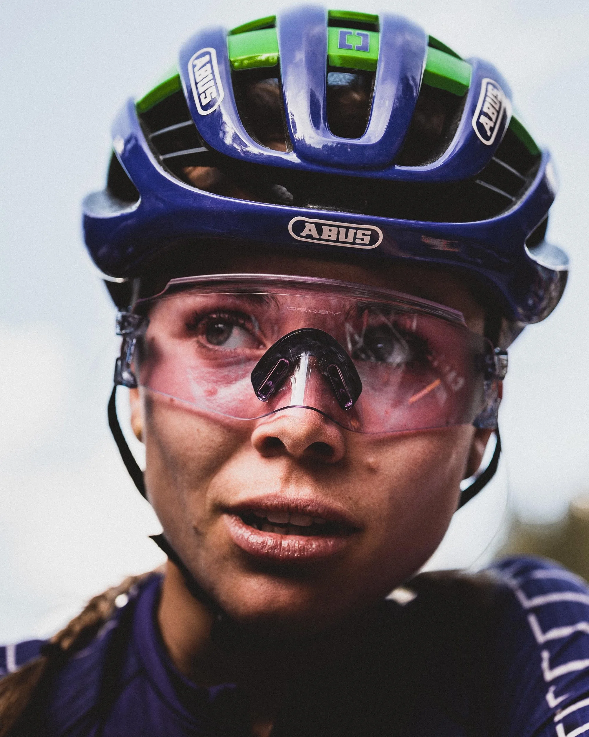 A female cyclist wearing a blue helmet with green accents, pink-tinted sunglasses, and a blue cycling jersey.