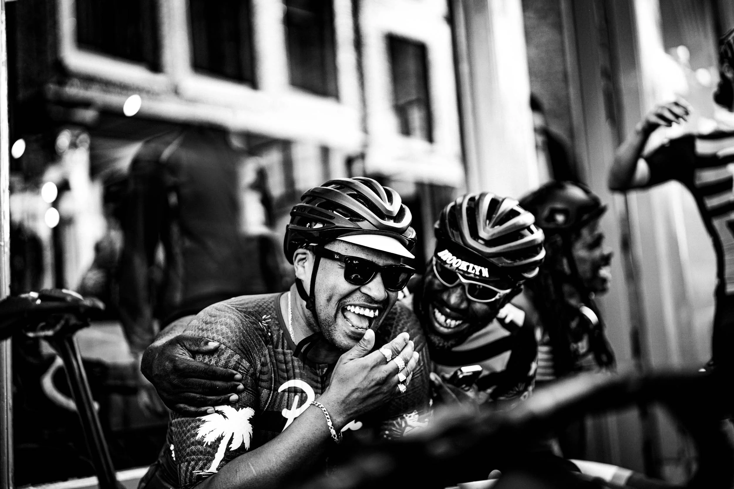 Two smiling men wearing helmets and sunglasses, sharing a joyful moment, with people and buildings in the background.