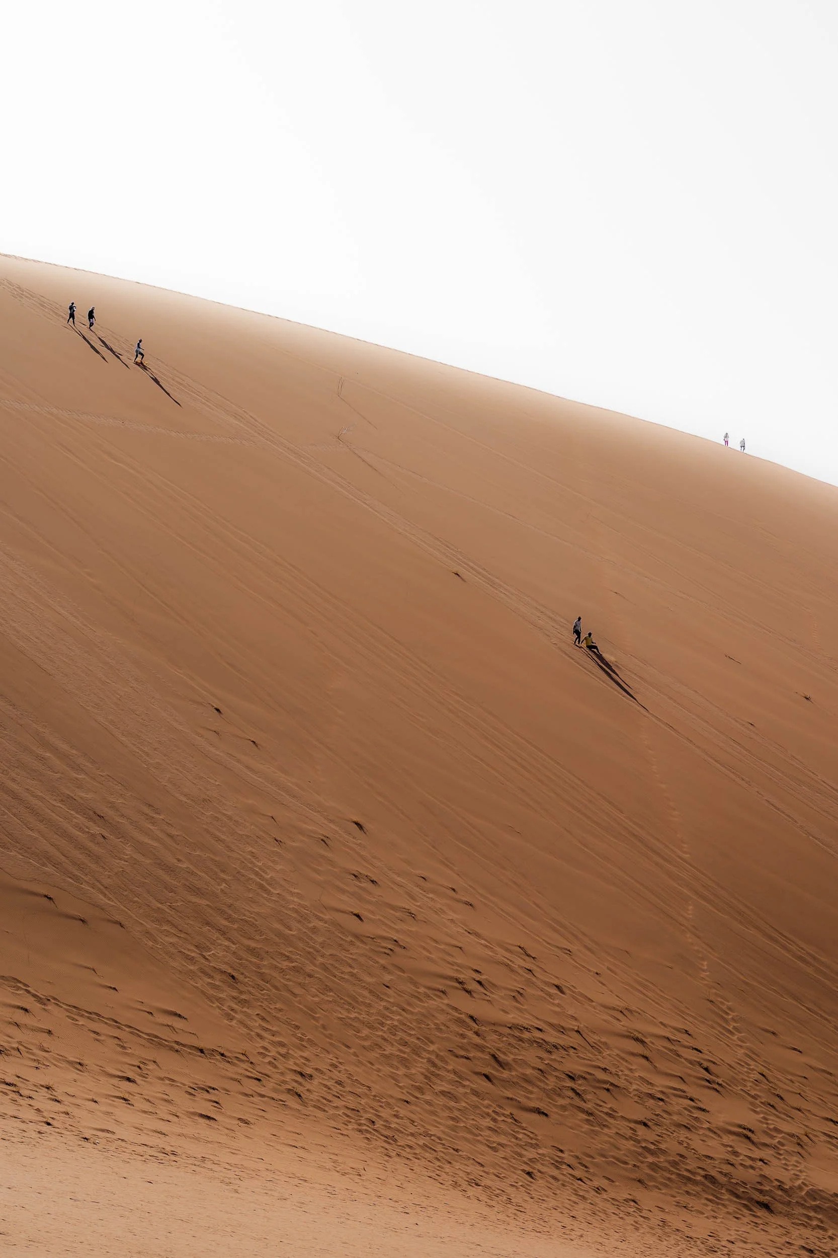 Multiple people hiking up sand dunes in a desert with a clear white sky.