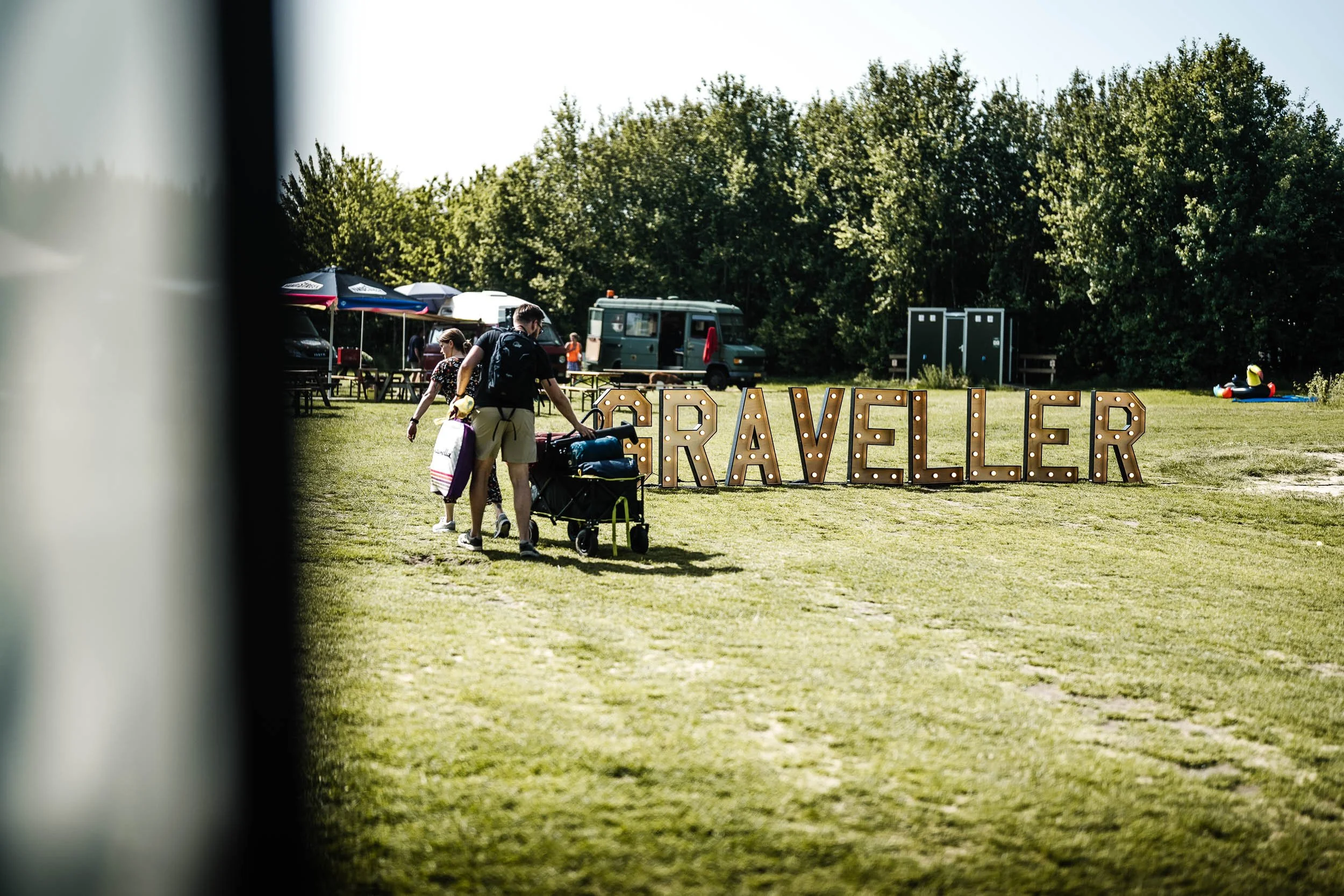 People walking towards large illuminated sign that spells out "TRAVELER" in an open grassy area with tents and trees in the background. (Graveller)