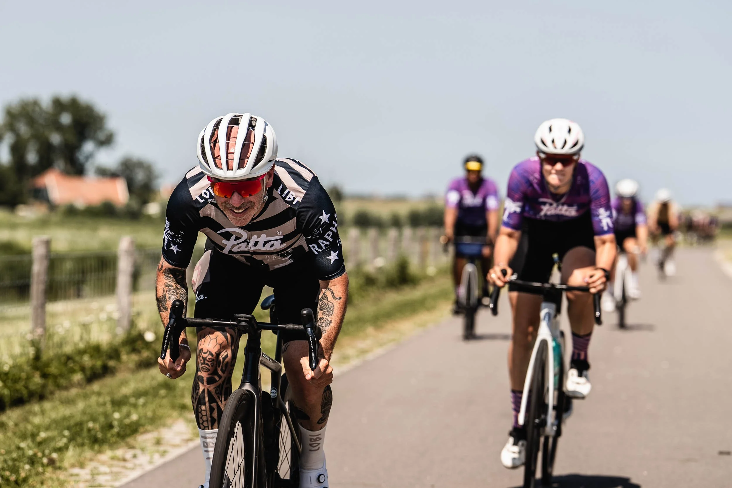 Group of cyclists riding on a rural road on a sunny day, with fields and a house in the background.