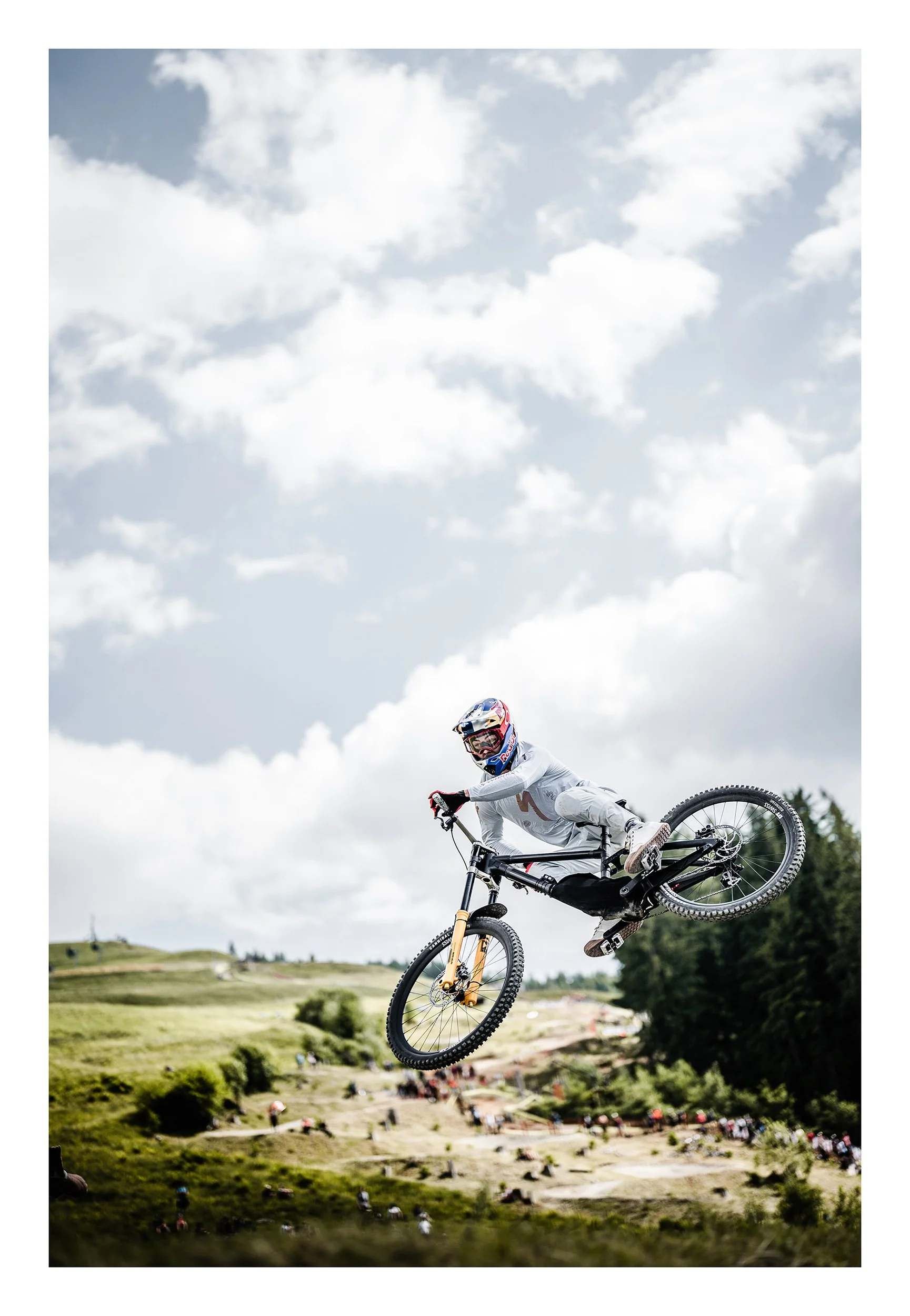 A mountain biker in a white outfit and helmet performing a jump with a full-suspension bicycle, over a hilly green landscape with spectators below, under a cloudy sky.