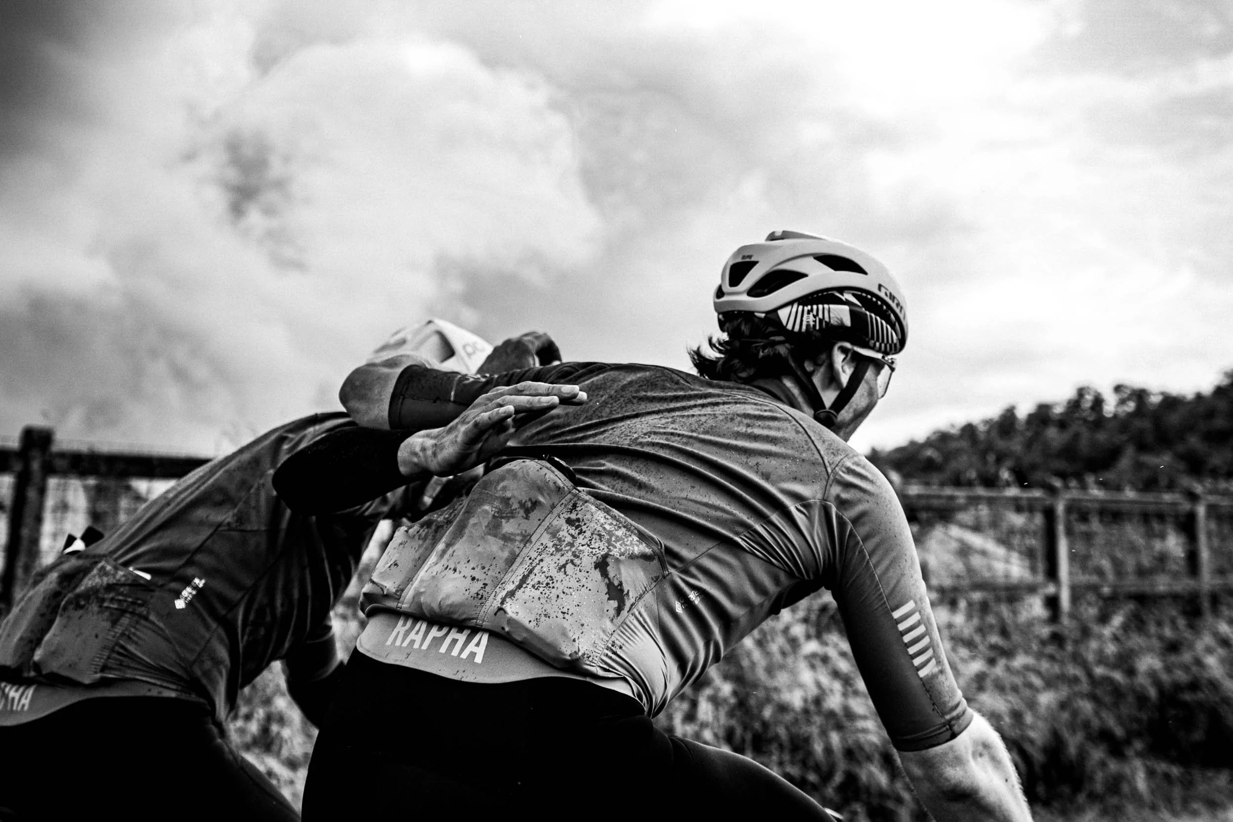 Two cyclists wearing helmets, one helping the other, on a dirt trail outdoors with cloudy sky and a fence in the background.