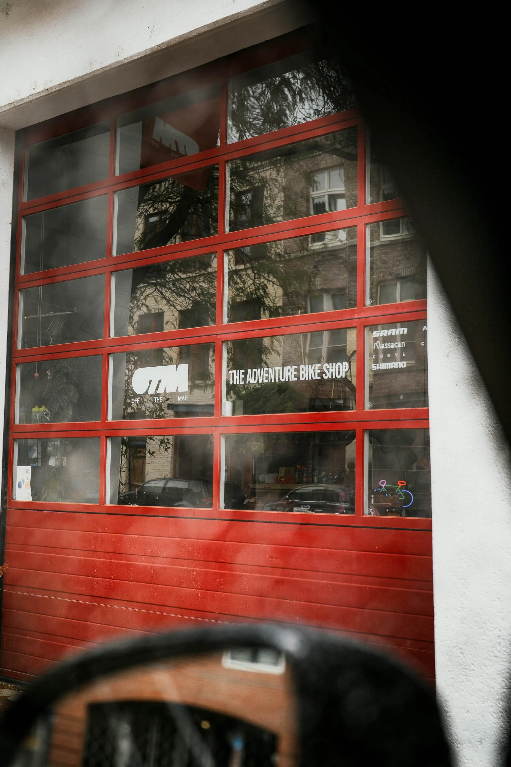 View of a garage door with large glass windows, signage reading 'The Adventure Bike Shop,' and bicycle art in the window. Reflection of buildings and trees can be seen on the glass.