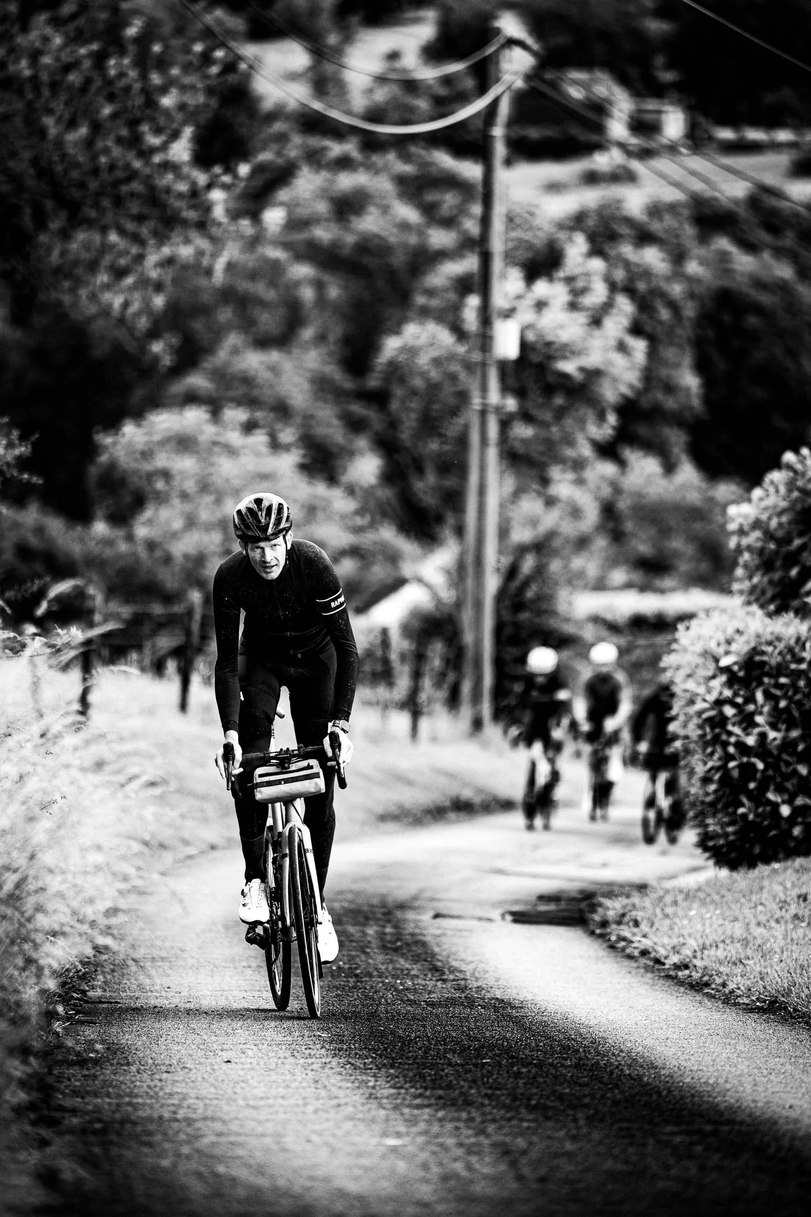 A cyclist riding uphill on a narrow road with a small group of cyclists blurred in the background, surrounded by trees and rural scenery.