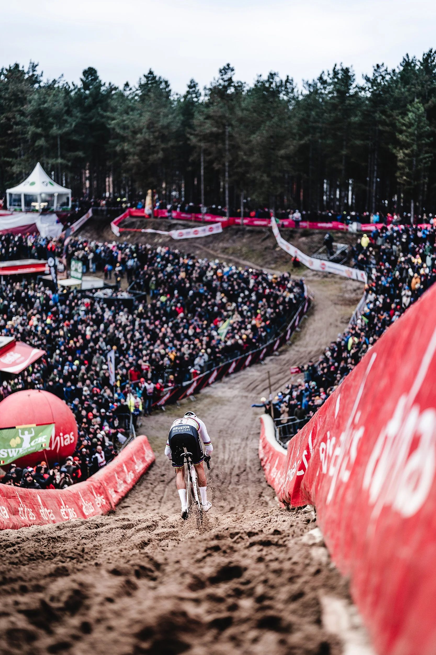 A cyclist riding on a dirt track during a race with a large crowd of spectators watching from the sides and a forest in the background.