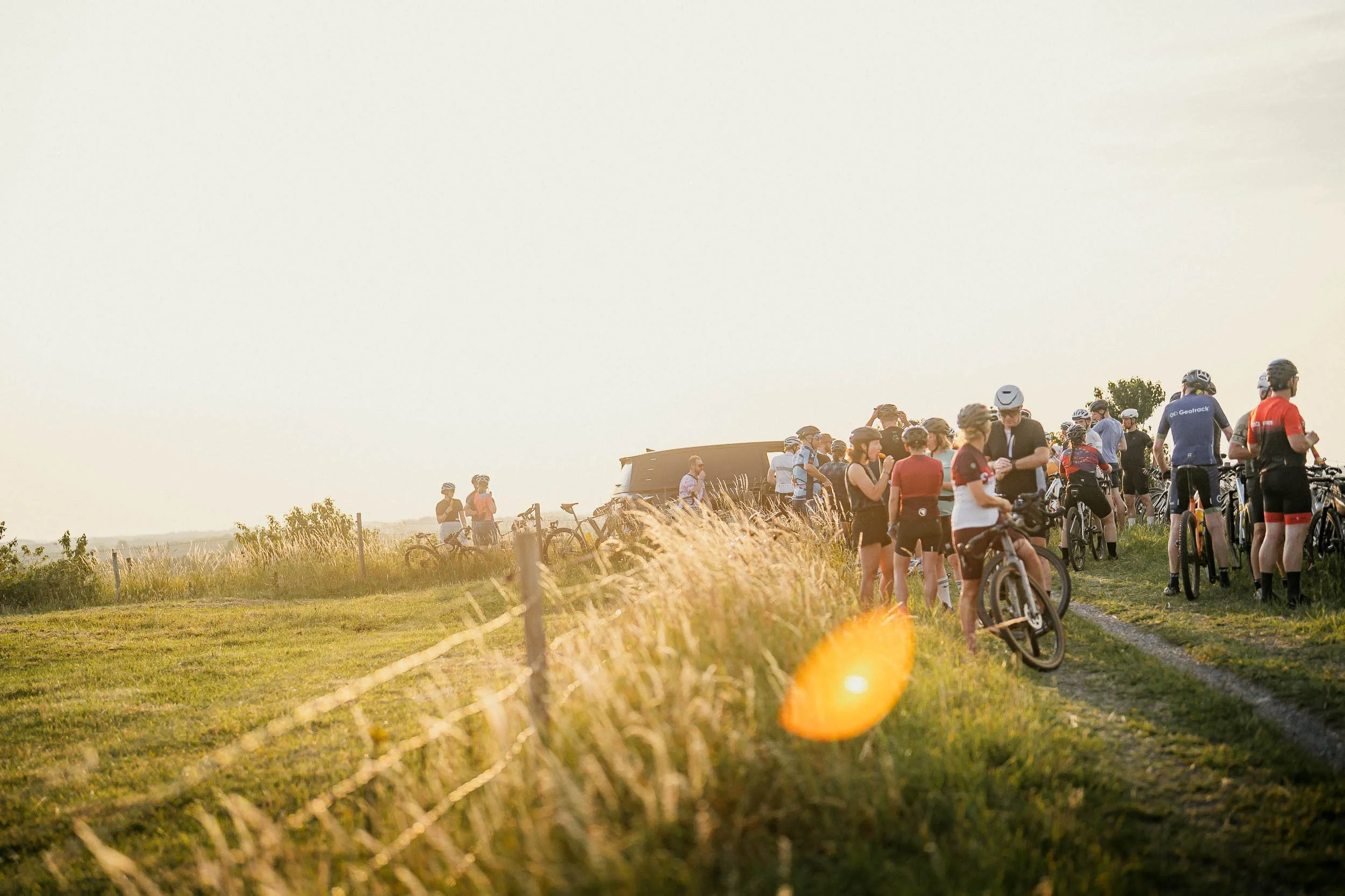 A group of cyclists and spectators gathering on a grassy field during sunset, with bicycles resting on the ground and people wearing helmets and cycling gear.(Graveller) )(Canyon)