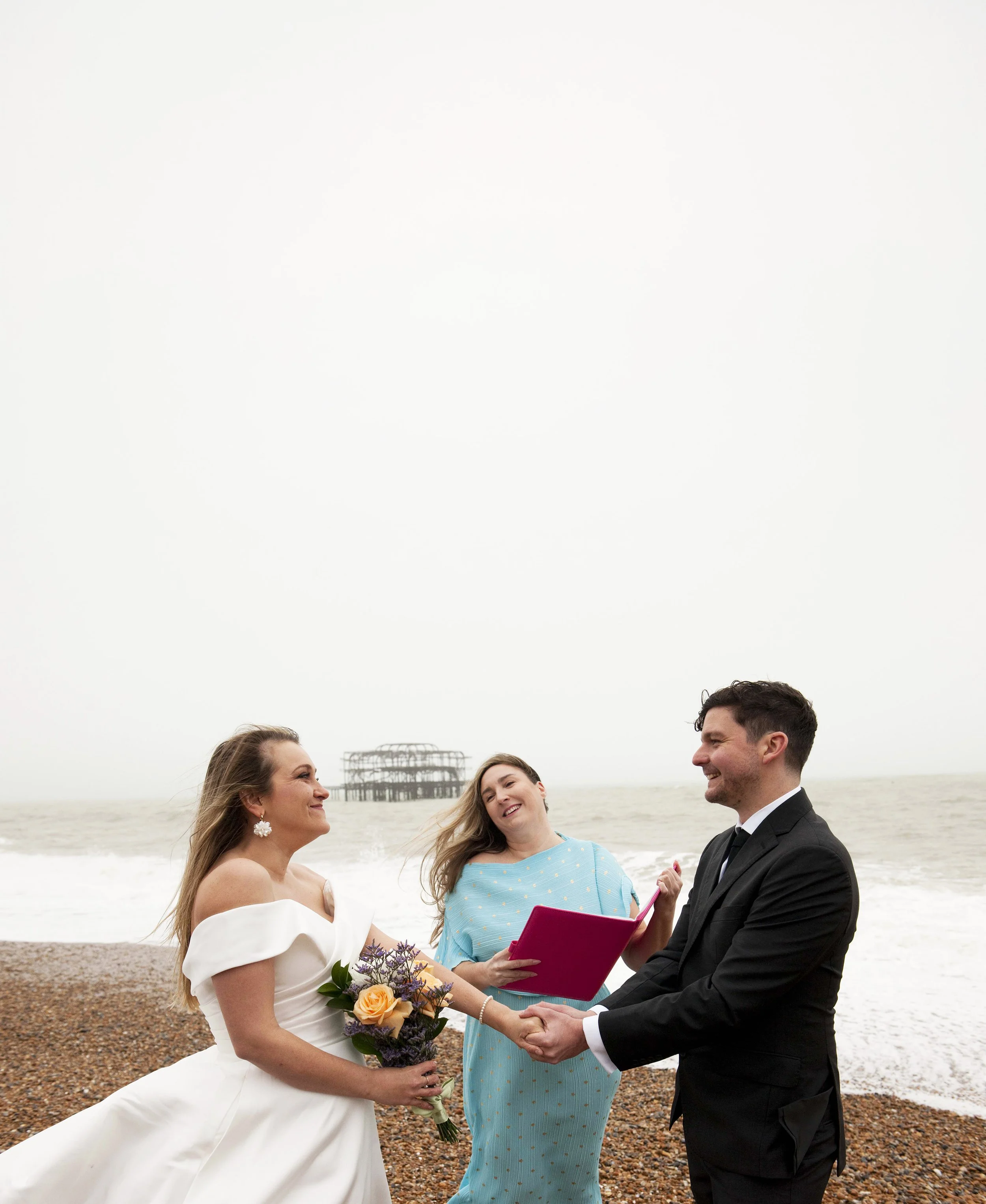 A wedding ceremony on a beach with a bride, groom, and two women. The bride is holding a bouquet of flowers and wearing a white wedding dress. The groom is in a black tuxedo. A woman in a light blue dress is officiating, holding a pink folder. The ot