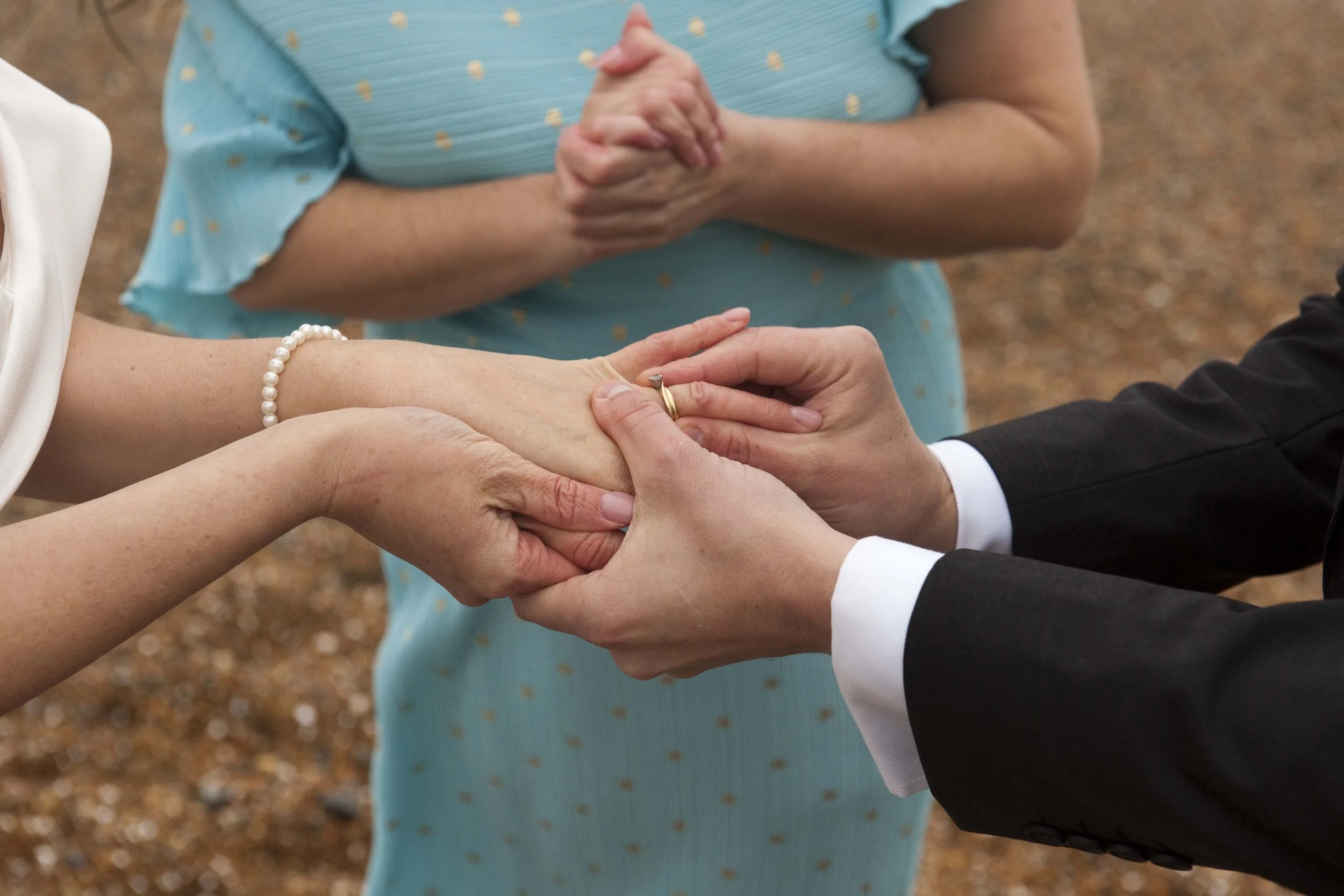 Person in a black suit holding hands in a wedding ceremony with another person wearing a pearl bracelet and ring, background out of focus.