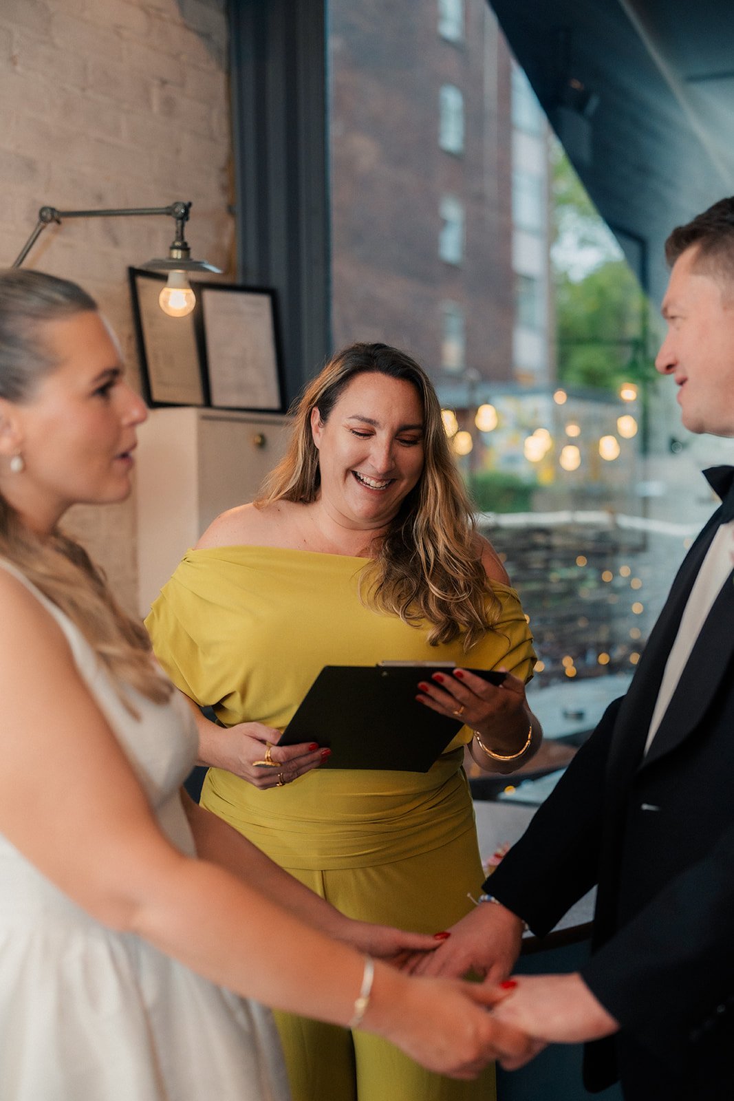 A couple getting married at a wedding ceremony, holding hands and looking at each other, with an officiant smiling while reading vows or an oath, inside a modern venue with brick walls and large windows.