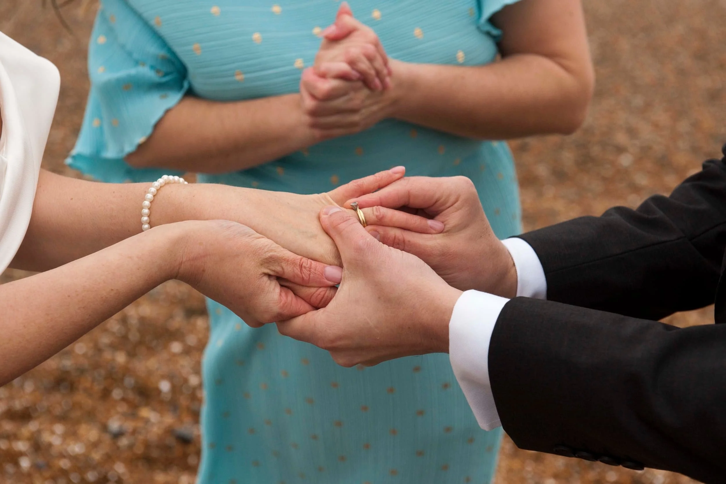 Person in a black suit holding hands in a wedding ceremony with another person wearing a pearl bracelet and ring, background out of focus.