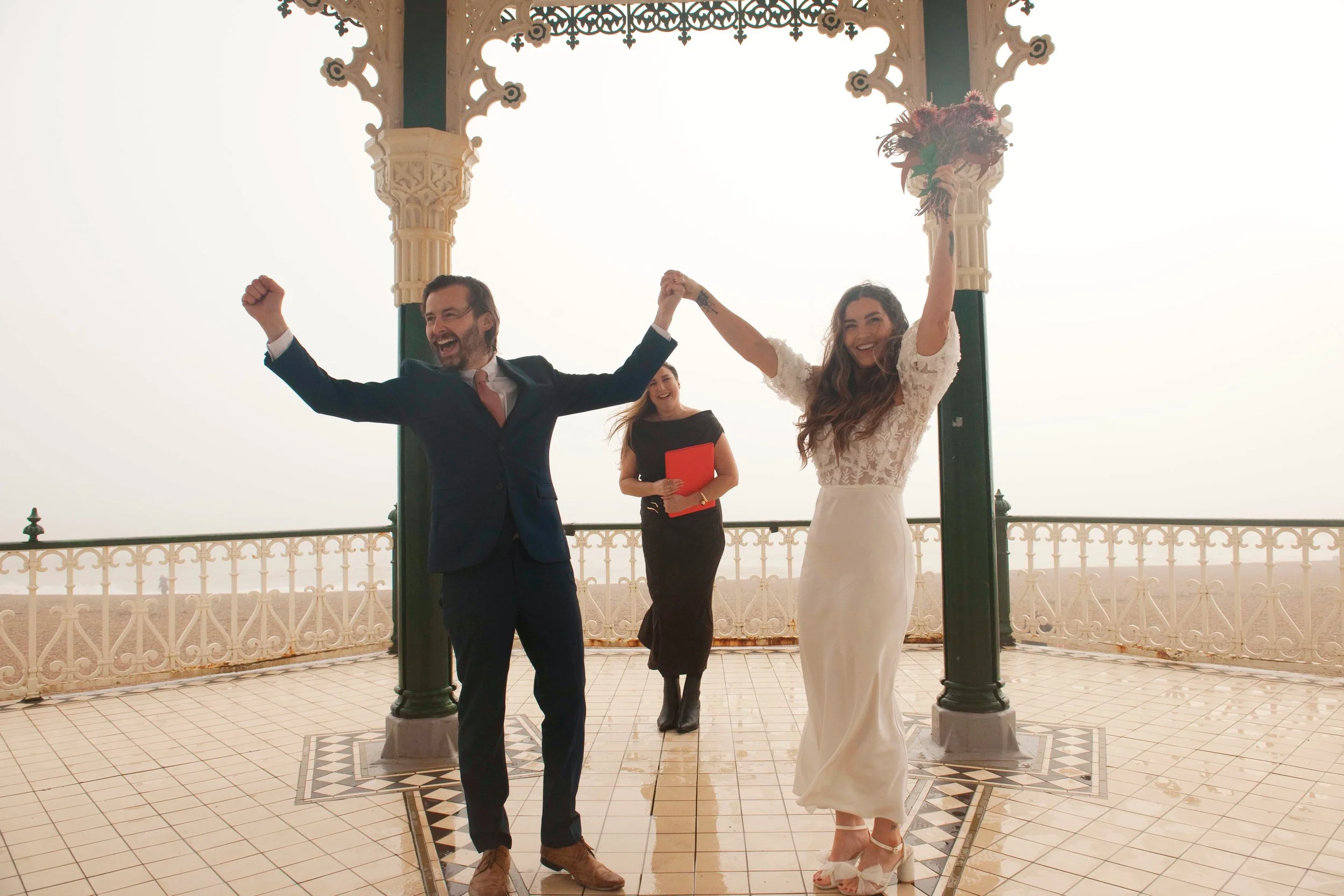 A joyful wedding scene with a man and woman holding hands and celebrating, standing under decorative green and cream pergola columns on a patterned tiled floor with ornate white railing, a woman in a black dress holding a red folder standing behind t
