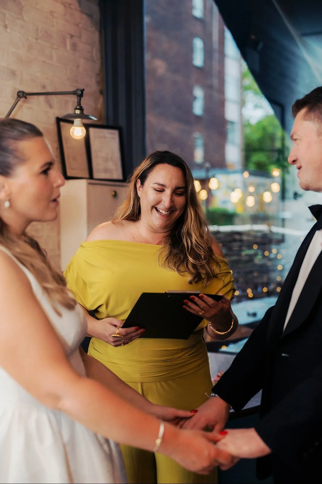 A couple getting married at a wedding ceremony, holding hands and looking at each other, with an officiant smiling while reading vows or an oath, inside a modern venue with brick walls and large windows.