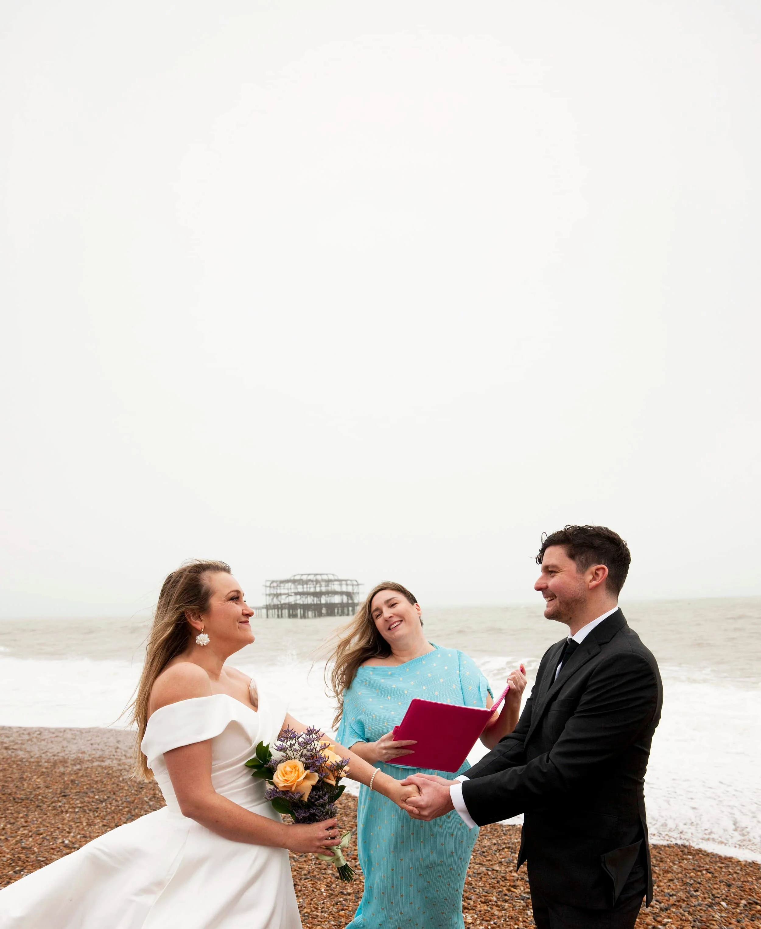 A wedding ceremony on a beach with a bride, groom, and two women. The bride is holding a bouquet of flowers and wearing a white wedding dress. The groom is in a black tuxedo. A woman in a light blue dress is officiating, holding a pink folder. The ot