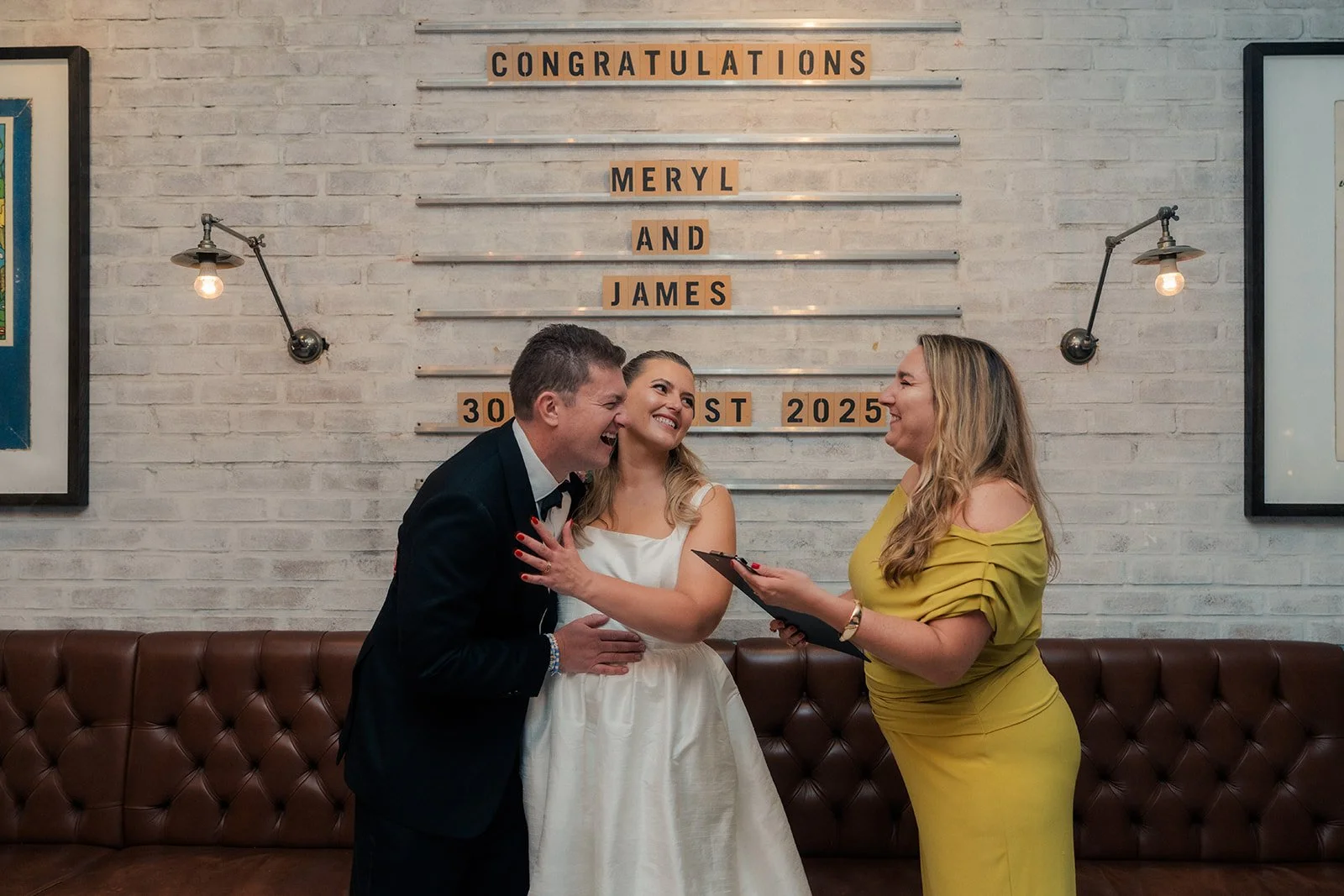 A wedding ceremony with a bride, groom, and officiant in front of a decorative white brick wall with a sign that reads 'Congratulations Meryl and James 30th August 2025.'