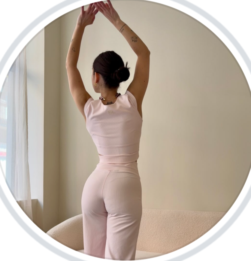 Mujer viste ropa de tono rosa claro, de espaldas, con cabello recogido en un moño, ajustándose la ropa con las manos levantadas, en un interior con luz natural y cortinas blancas.