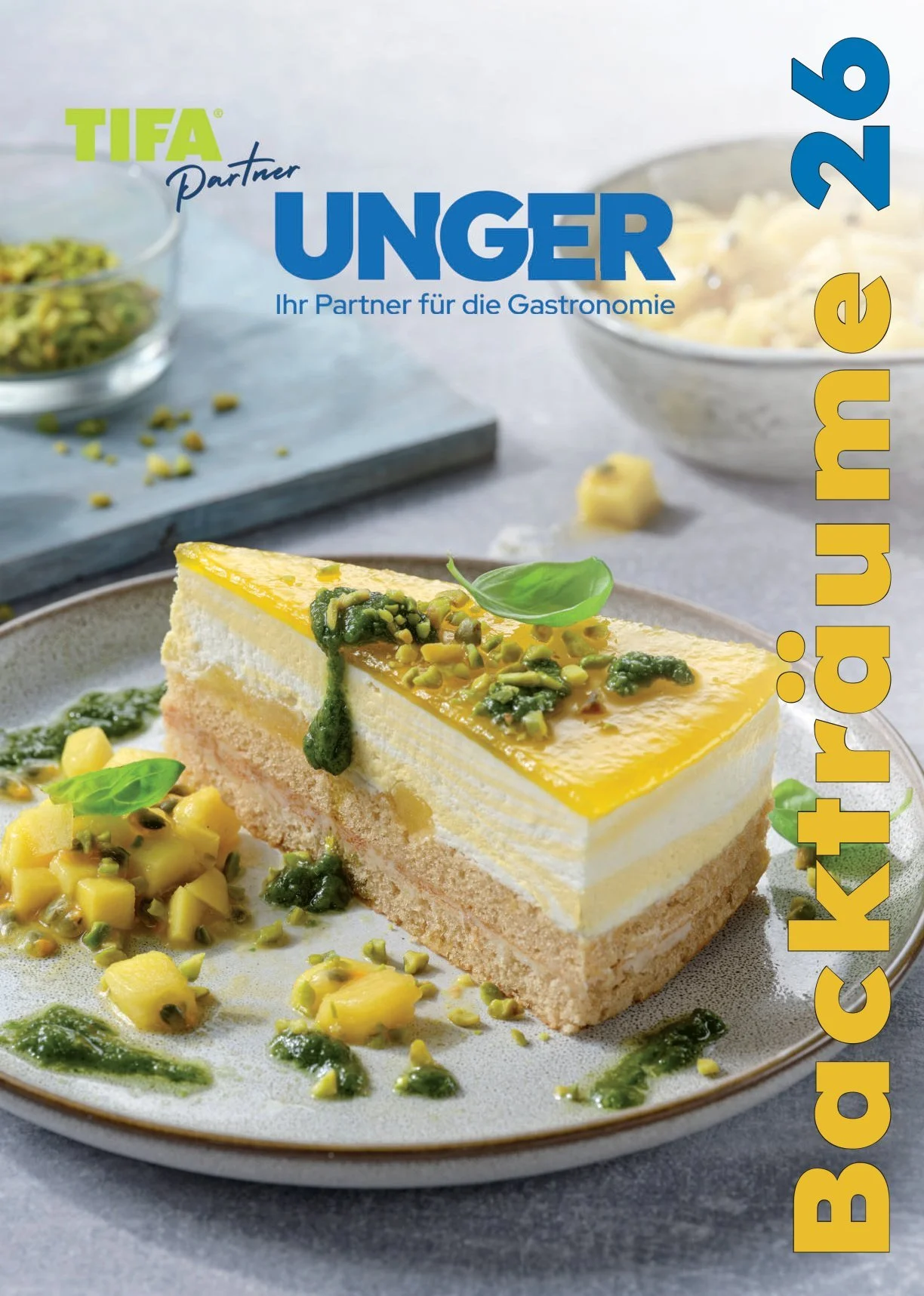 Buchcover eines Kochbuchs mit einer Torte, gelber und grüner Dekoration, auf einem Teller serviert, mit weiteren Zutaten im Hintergrund. Das Buch trägt den Titel 'Backträume 26'.