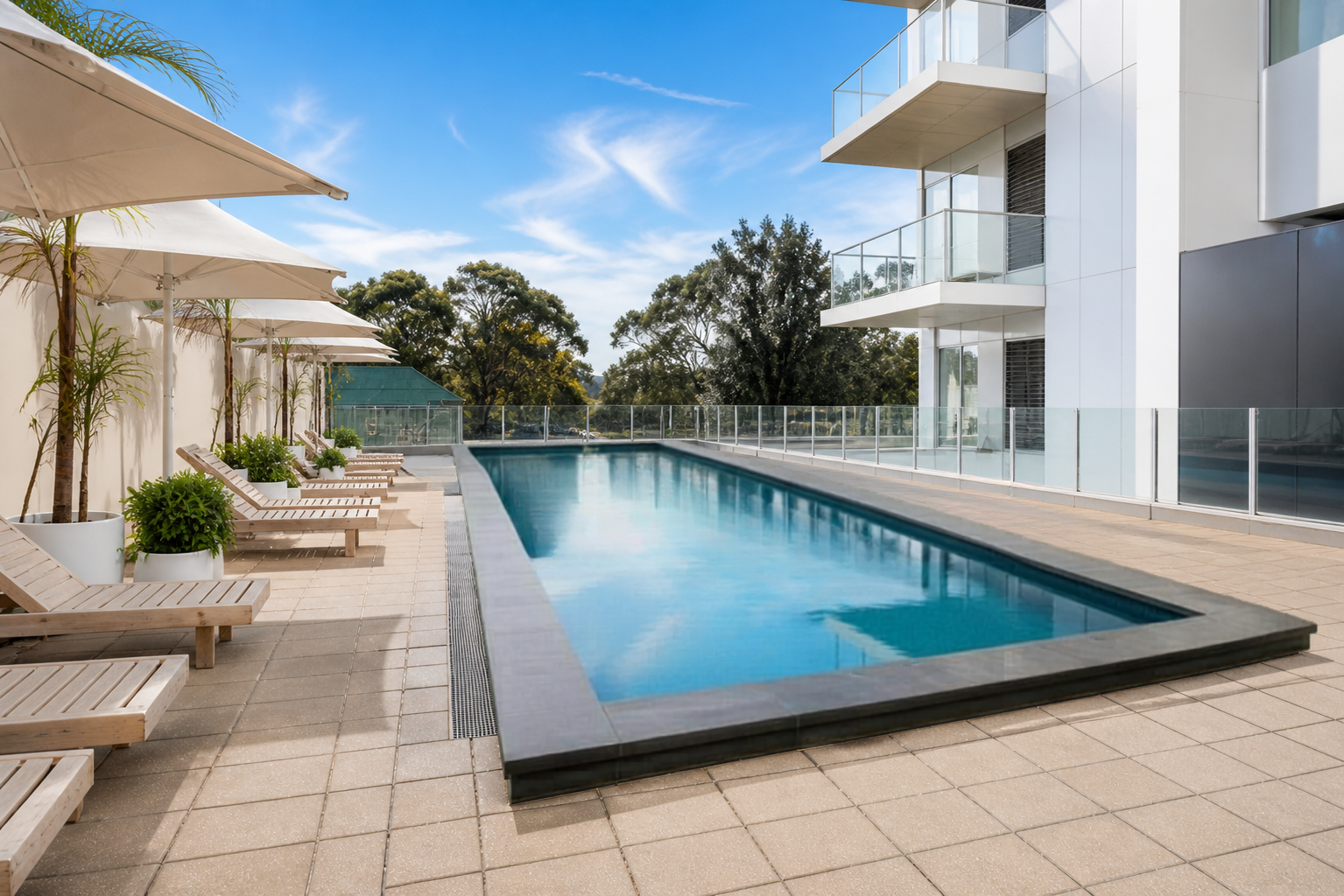 Modern residential outdoor swimming pool with sun loungers, umbrellas, potted plants on a tiled deck, and a glass railing, adjacent to a white apartment building with balconies, under a blue sky with clouds and surrounded by green trees.