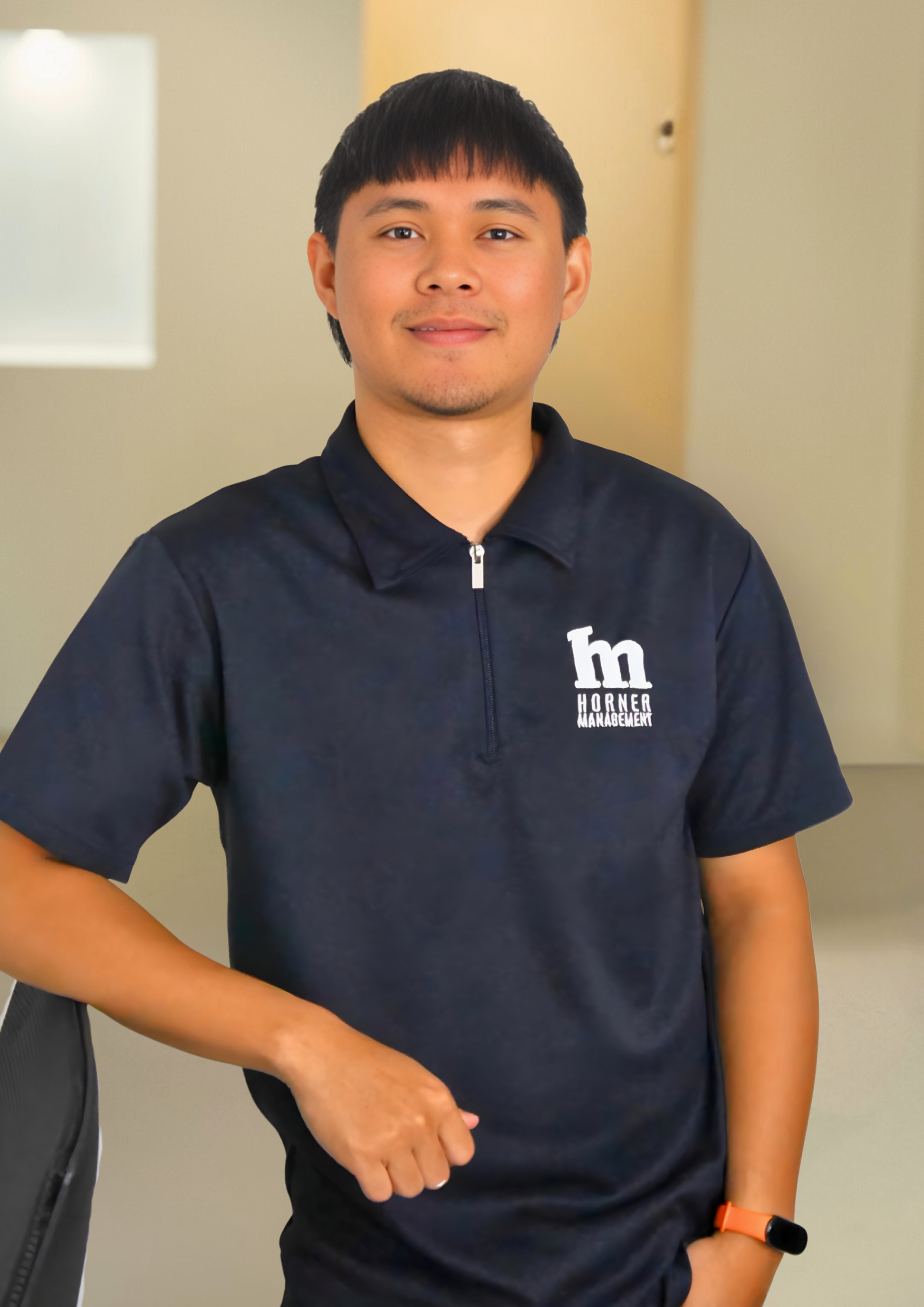 Young man with black hair, wearing a navy blue polo shirt with a logo on the chest, sitting indoors with a beige wall and door in the background, wearing an orange watch on his left wrist, smiling softly at the camera.