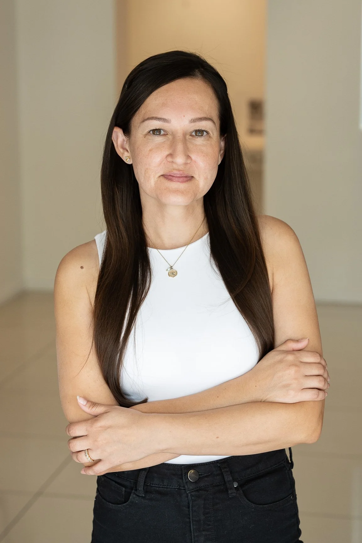 A woman with long brown hair, wearing a white sleeveless top and black jeans, standing with her arms crossed in a well-lit indoor space.
