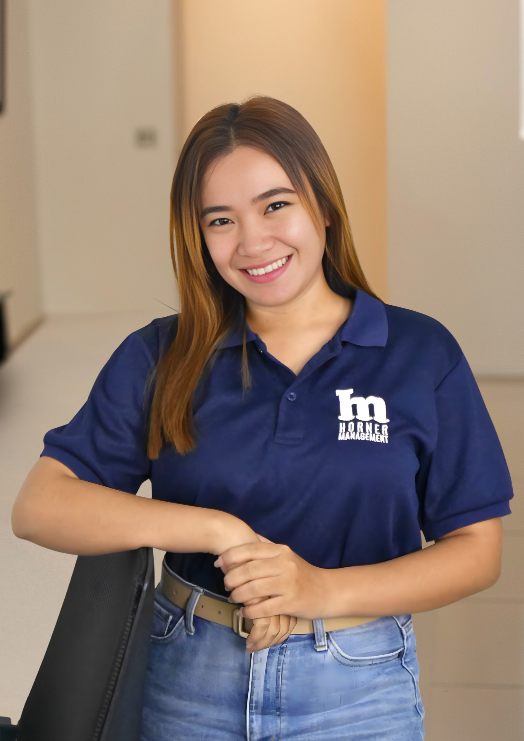 Young woman with long brown hair smiling, wearing a navy blue polo shirt with a logo and text, and blue jeans, sitting at a desk with a black chair.
