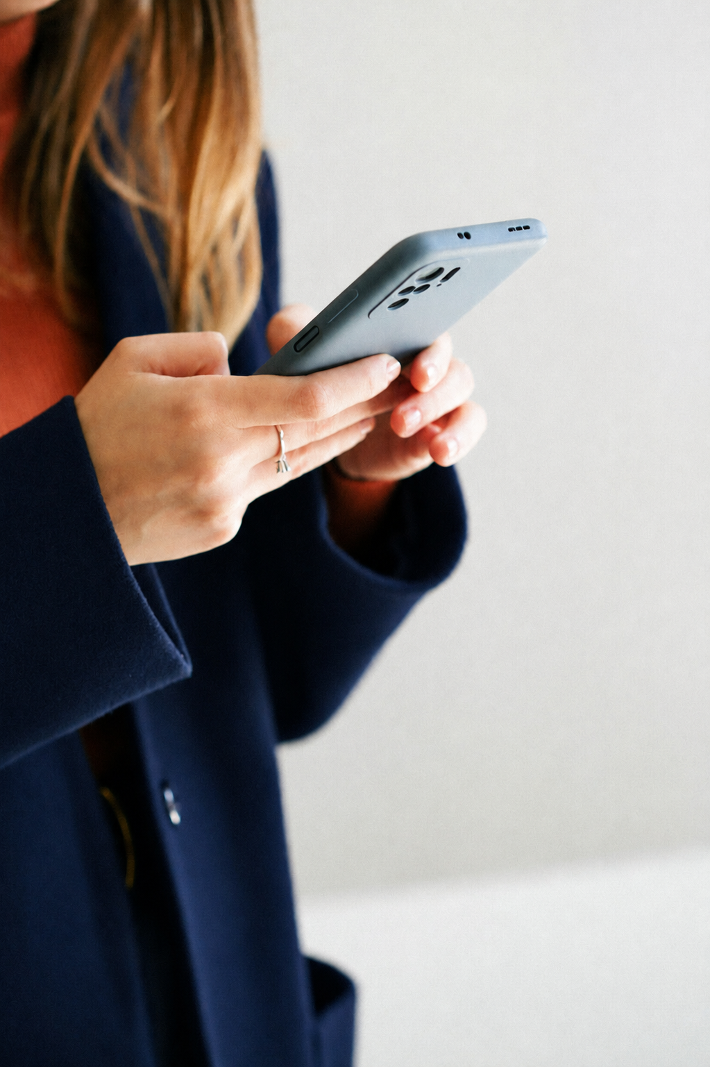 A woman with long hair wearing a navy jacket and rust-colored top, holding and looking at a smartphone.