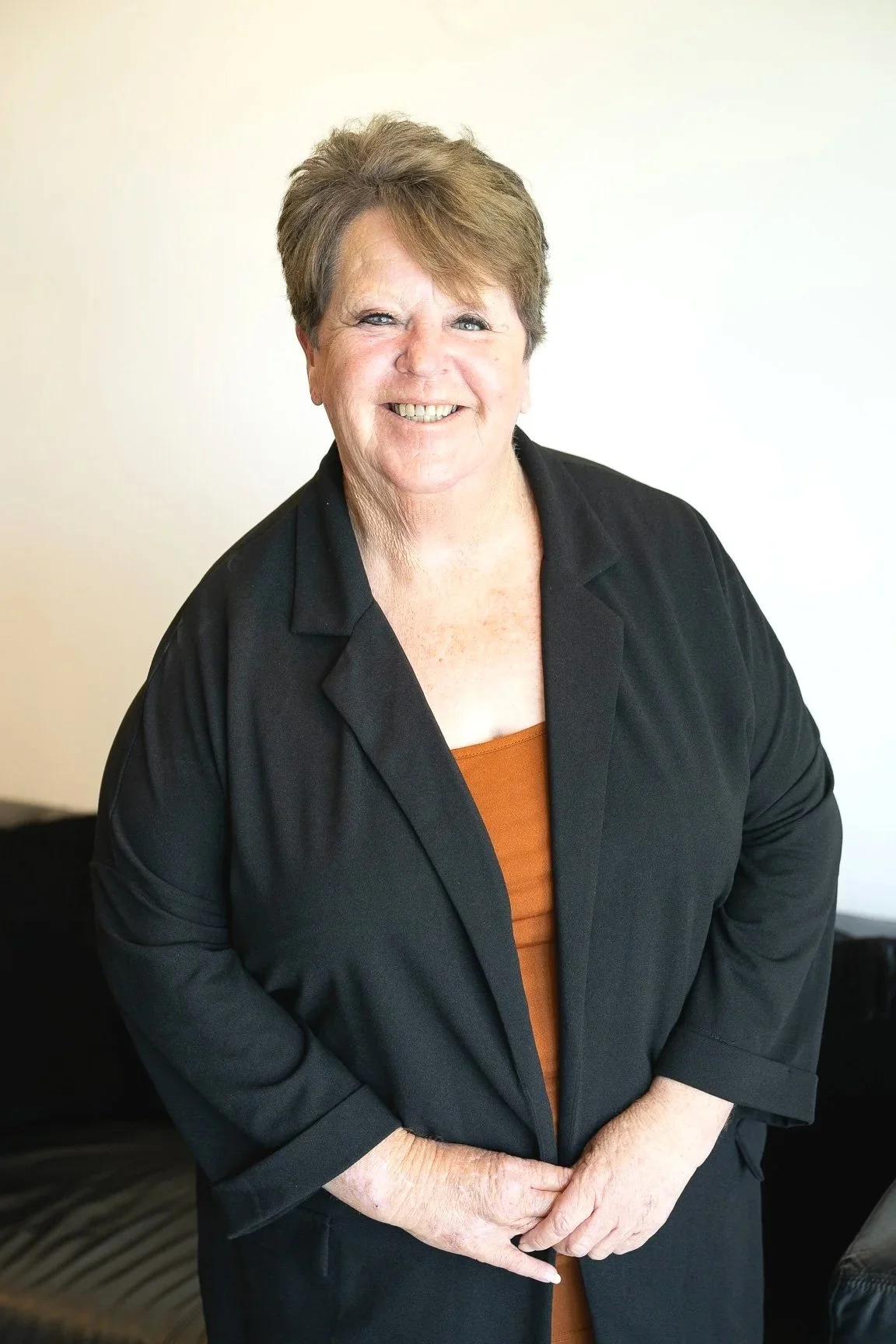 A smiling older woman with short light brown hair, wearing a black blazer over a rust-colored top, standing against a plain white wall.