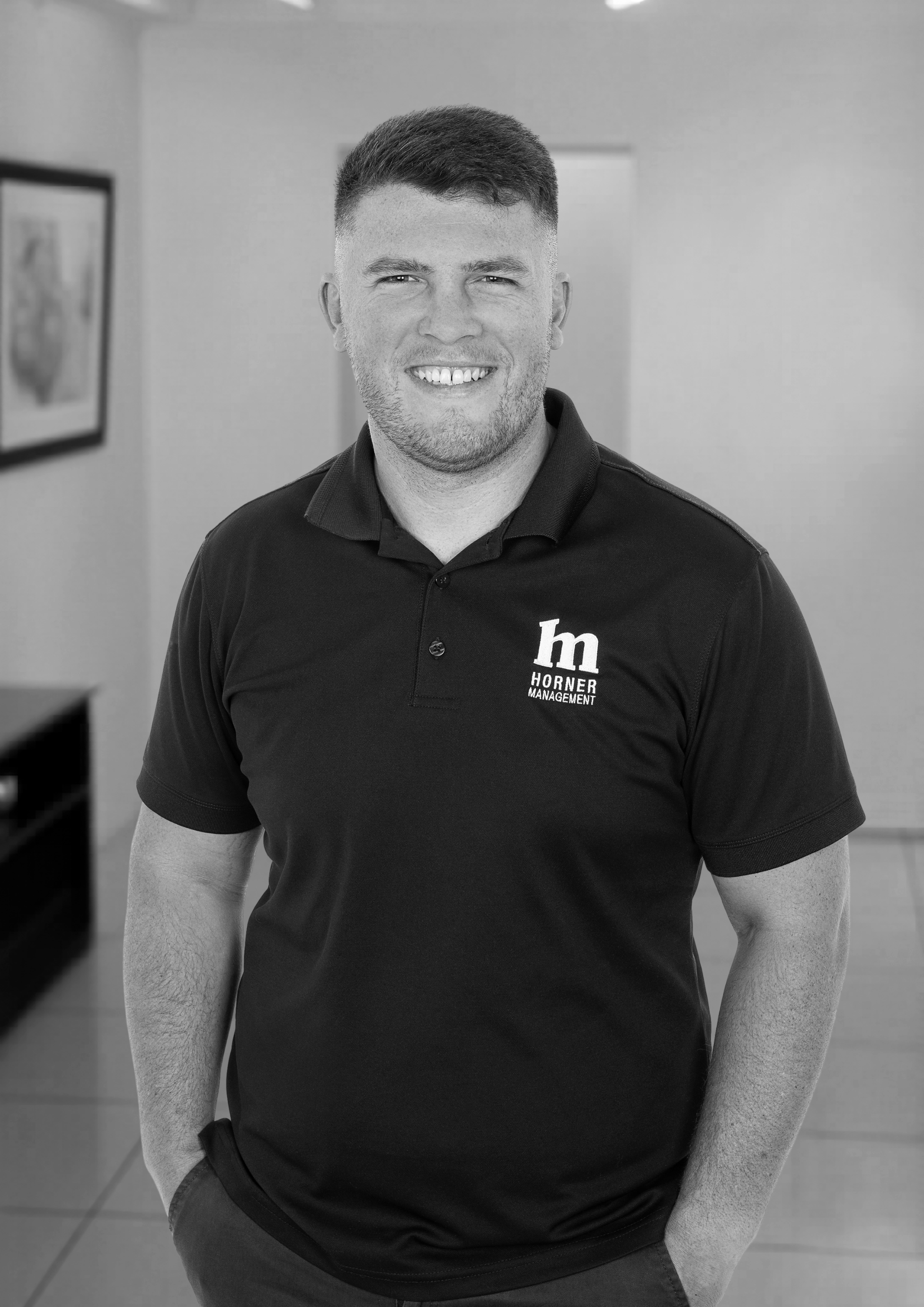 Black and white photo of a smiling Luke Scicluna, General Manager of Horner Management. He is standing indoors with a framed picture on the wall behind him.