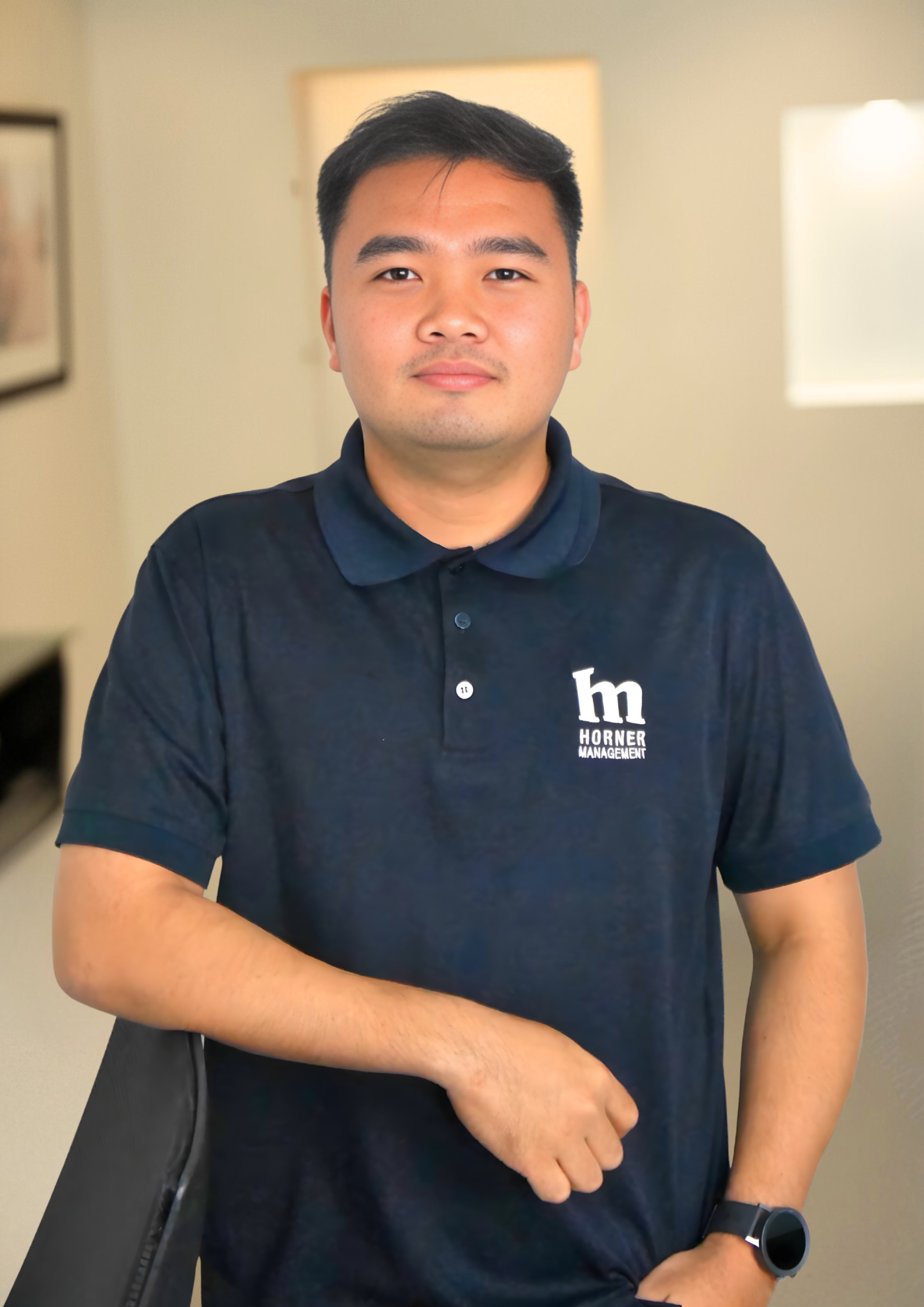 A young man with short black hair and a light complexion standing indoors. He is wearing a dark blue polo shirt with a logo that reads 'h m HORNER MANAGEMENT' and a black wristwatch. The background includes blurred lights and artwork on the wall.