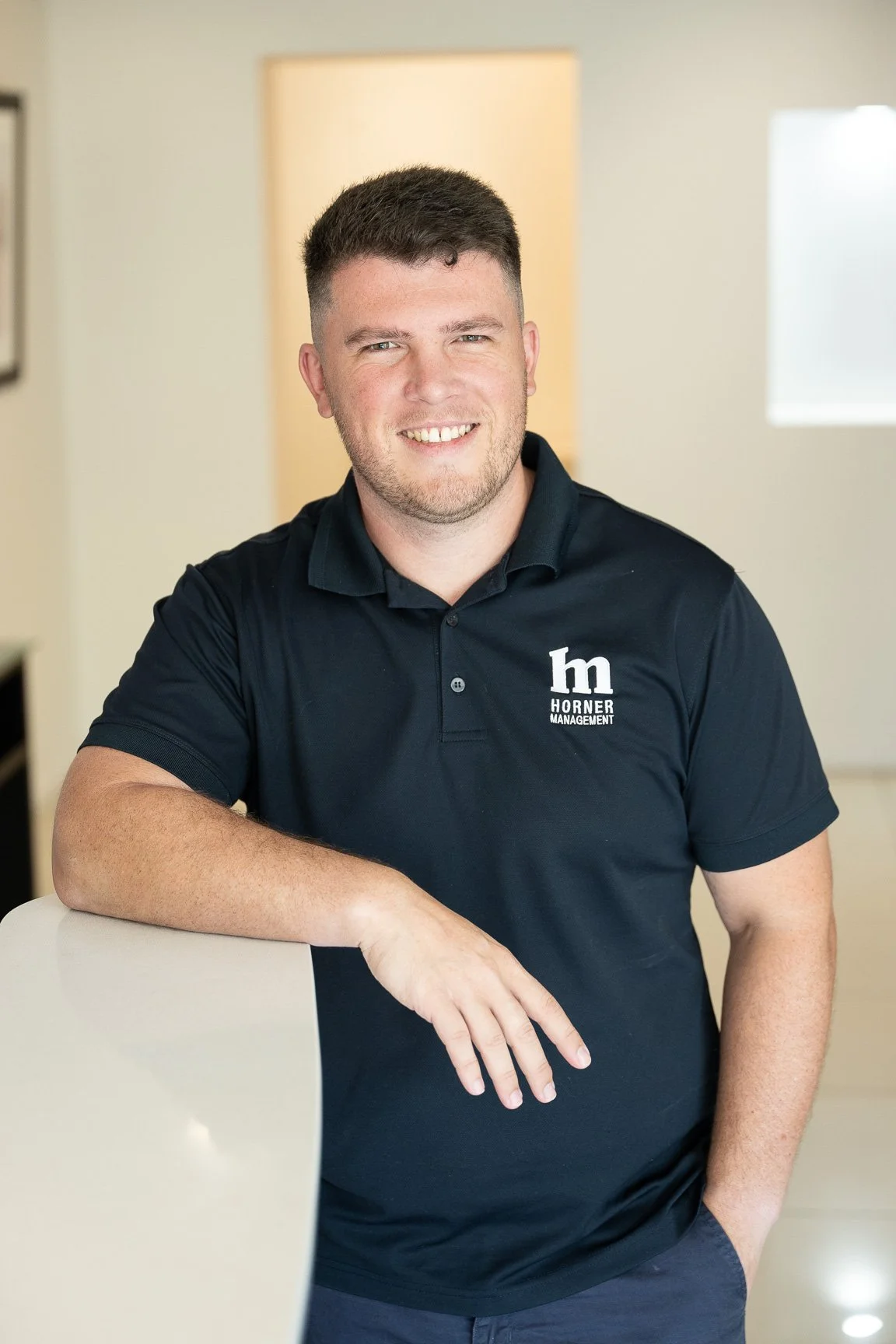 A young man with short brown hair and a trimmed beard, smiling, wearing a navy blue polo shirt with a logo that reads "h management" and the letter "m" on it. He is leaning on a white counter in an indoor setting with bright natural light.
