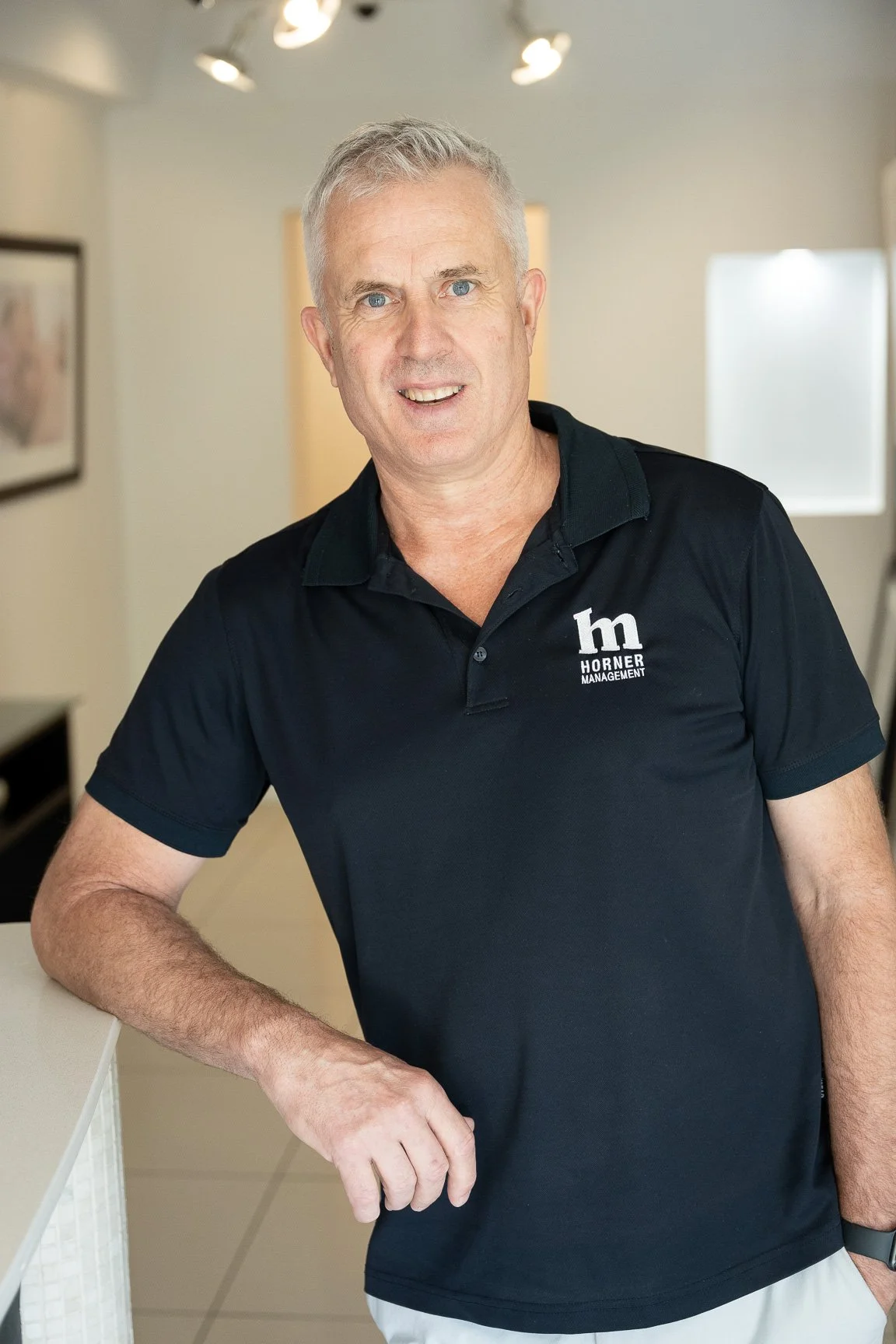 A middle-aged man with gray hair and blue eyes, wearing a black polo shirt with the logo 'Horner Management,' standing indoors, smiling at the camera.
