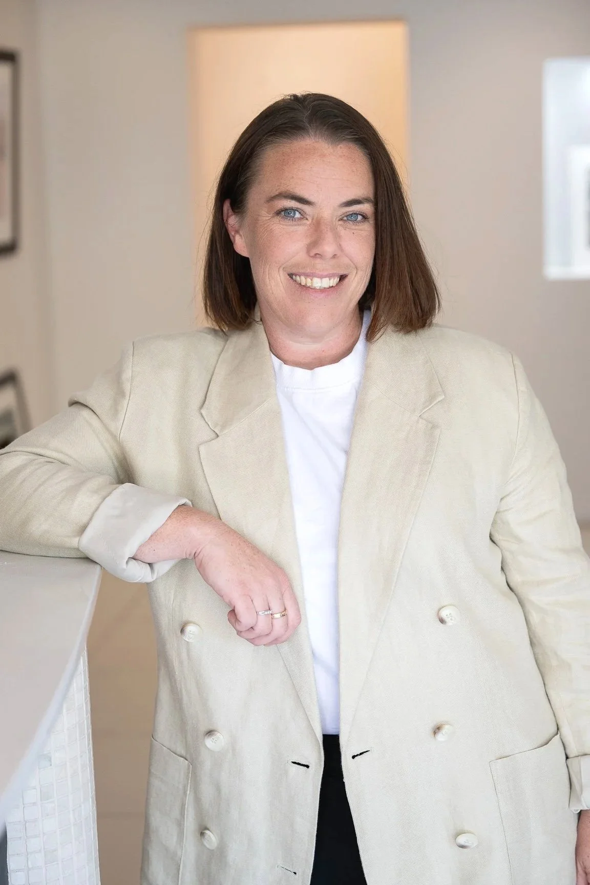 A woman with shoulder-length brown hair, blue eyes, and a bright smile, wearing a beige blazer over a white top, standing indoors with her arm resting on a white surface, in a well-lit room with a lamp and window in the background.