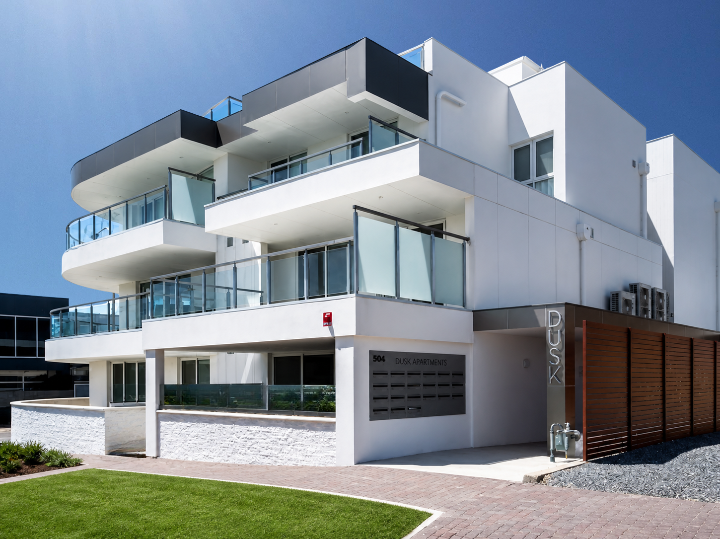 Modern white apartment building with glass balconies, including a sign showing that it is 'Dusk Apartments' in Henley Beach, it has a green lawn and a brick sidewalk in front.