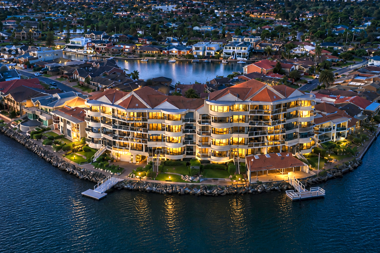 Aerial view of a waterfront residential complex at dusk. The complex features a multi-story building with illuminated interior lights, next to a lake with a dock and small boat on the edge. Surrounding the West Lakes complex are other houses.
