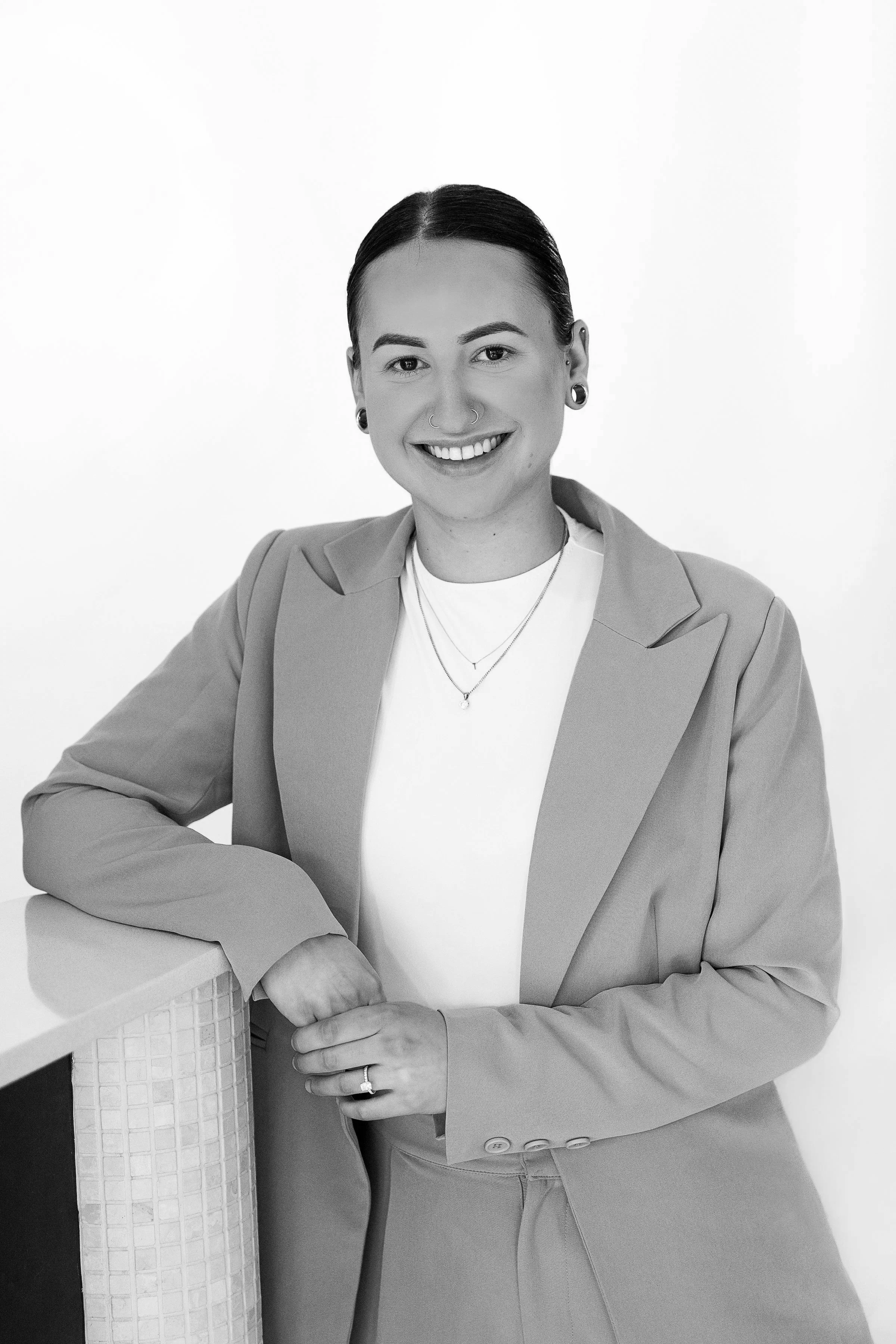 Black and white portrait of a young woman smiling, wearing a blazer, t-shirt, and layered necklaces, with her hair slicked back, standing next to a textured cylindrical object.